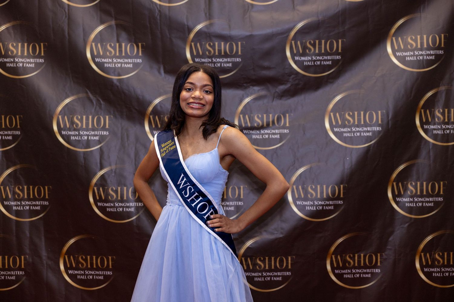 Young woman in blue gown wearing WSHOF sash poses confidently at Women's Humanitarian Hall of Fame event.