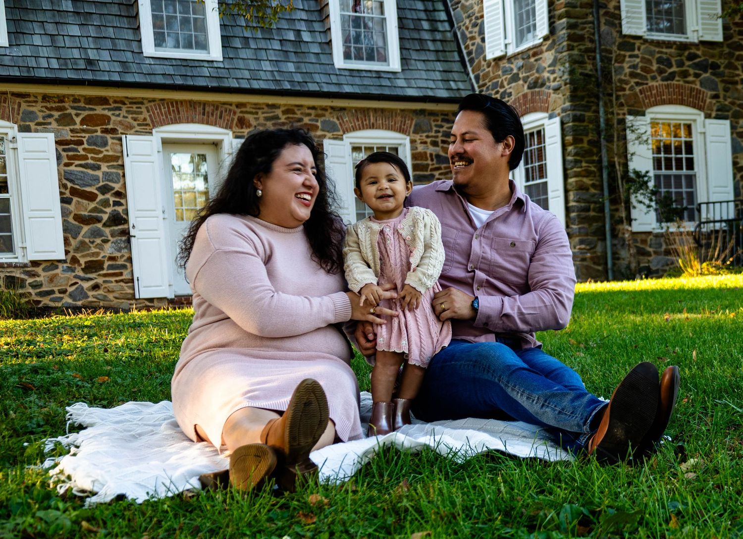 Happy family with toddler daughter sitting on blanket in grass outside stone house.