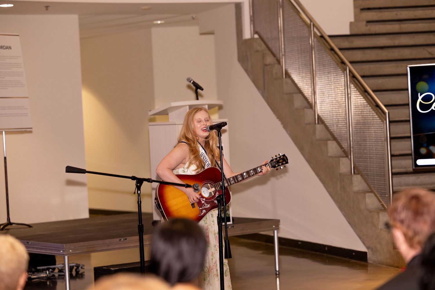 Female guitarist performs live on stage wearing a white gown at an indoor event.