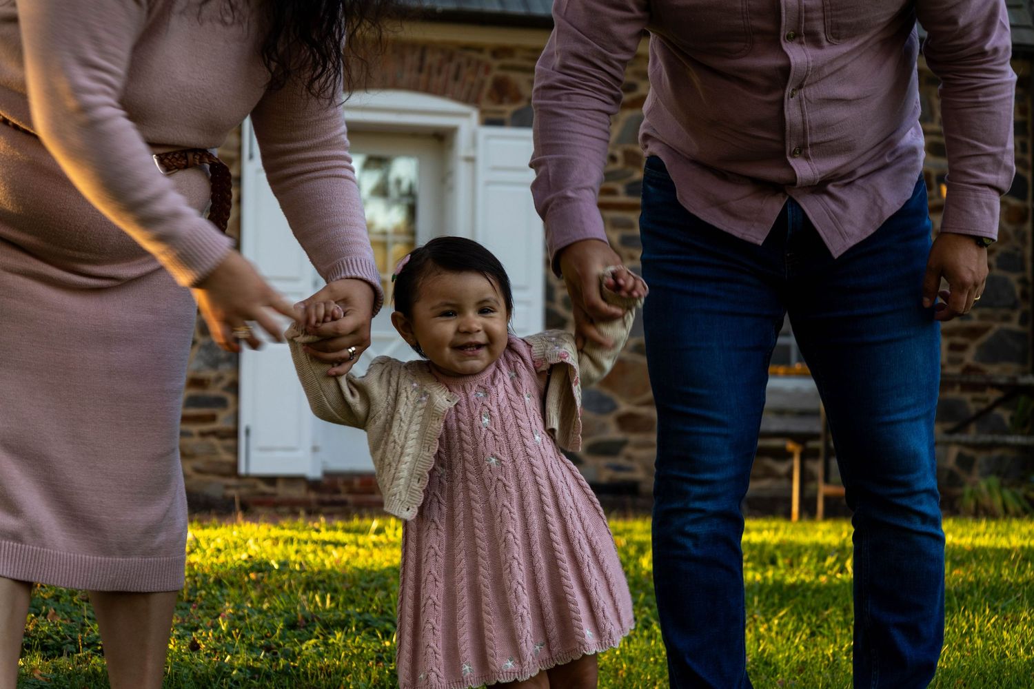 A joyful toddler girl in a pink dress walks outdoors holding hands with her parents on a sunny green lawn.