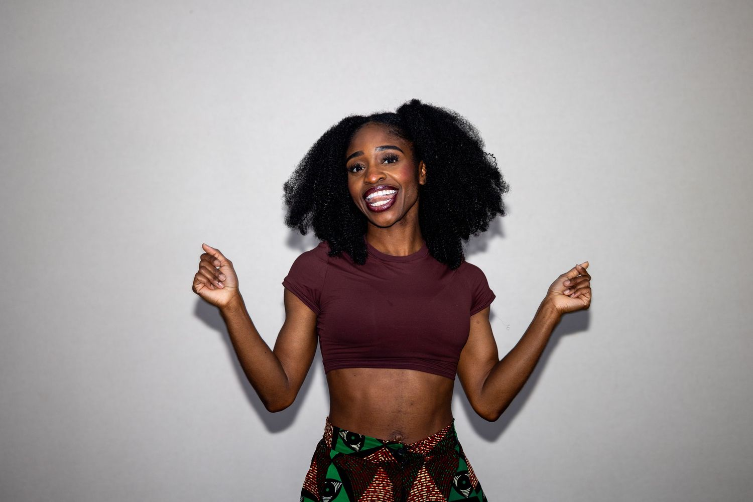 Joyful Black woman with natural afro hair wearing a maroon crop top and colorful printed skirt, dancing.
