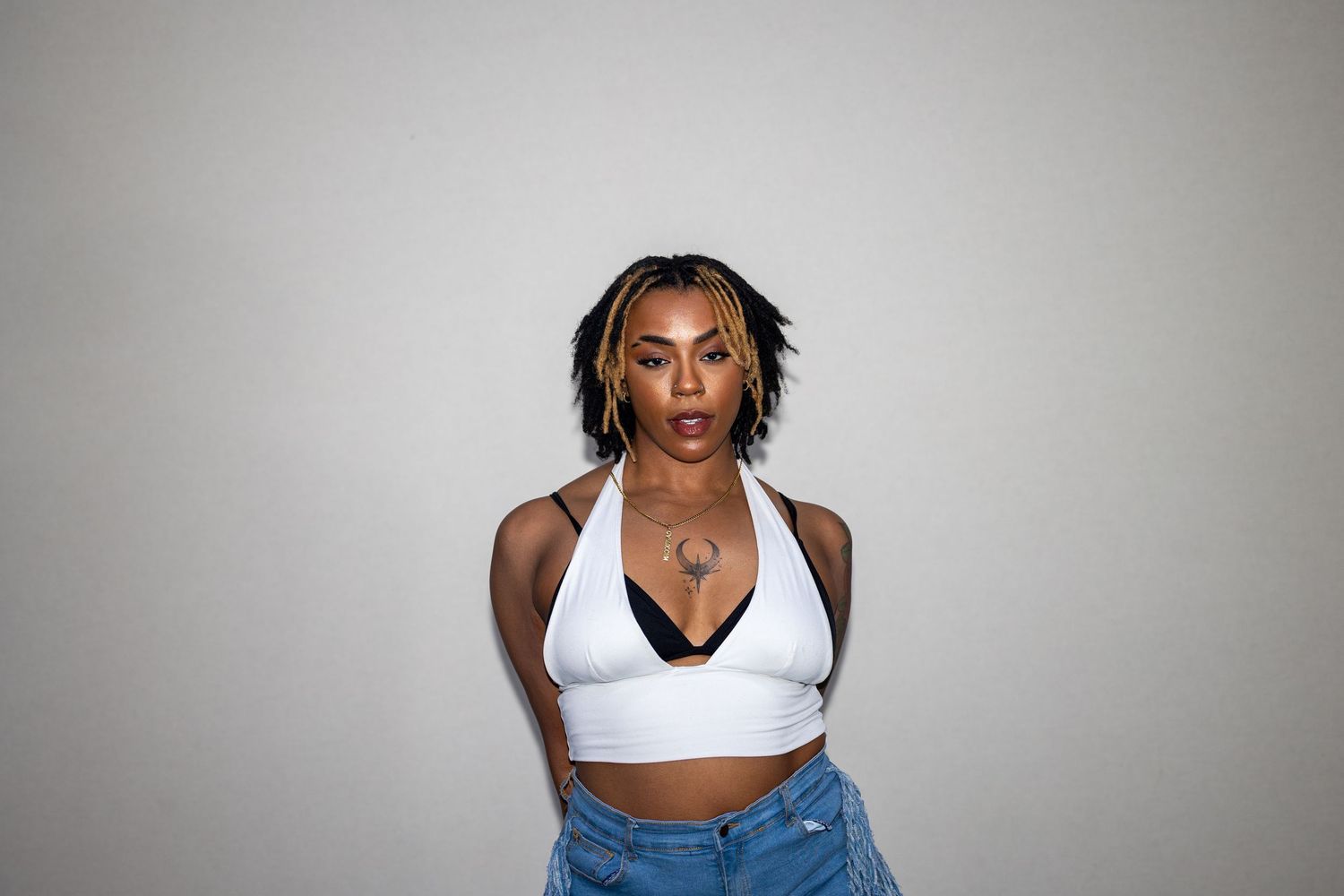 Young woman with braided hair wearing a white halter crop top and jeans posing against a gray background.