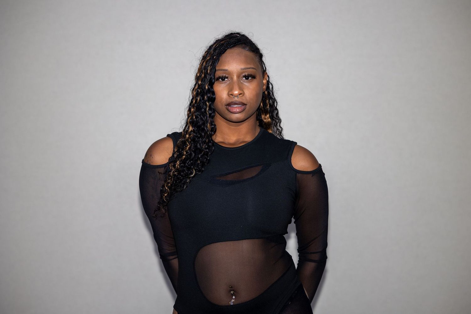 Woman in black cut-out top with braided hair poses confidently against a light gray background.