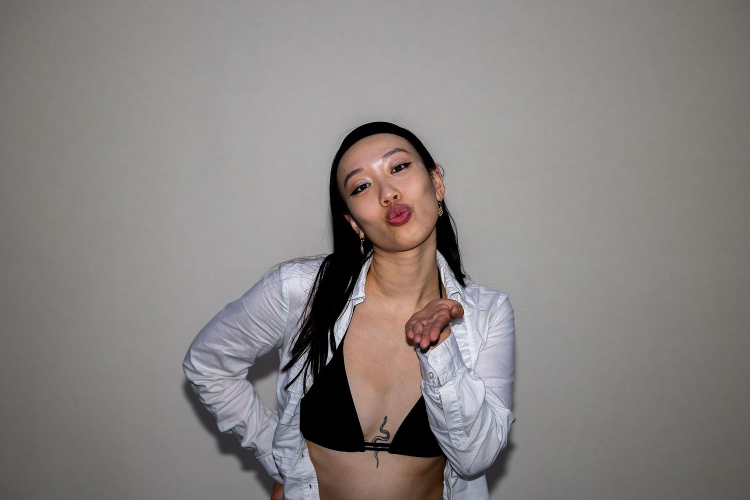 Young woman with dark hair wearing a white shirt and black bralette blowing a kiss against a gray background.