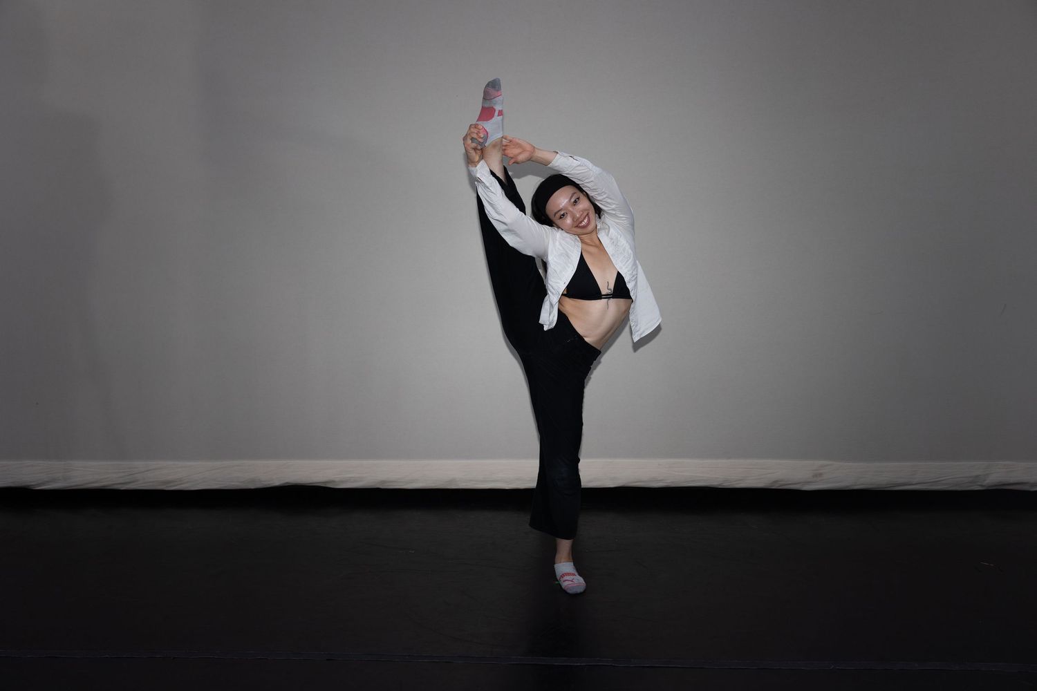 A dancer balances gracefully on one leg, extending the other high overhead while wearing black attire and a white jacket.