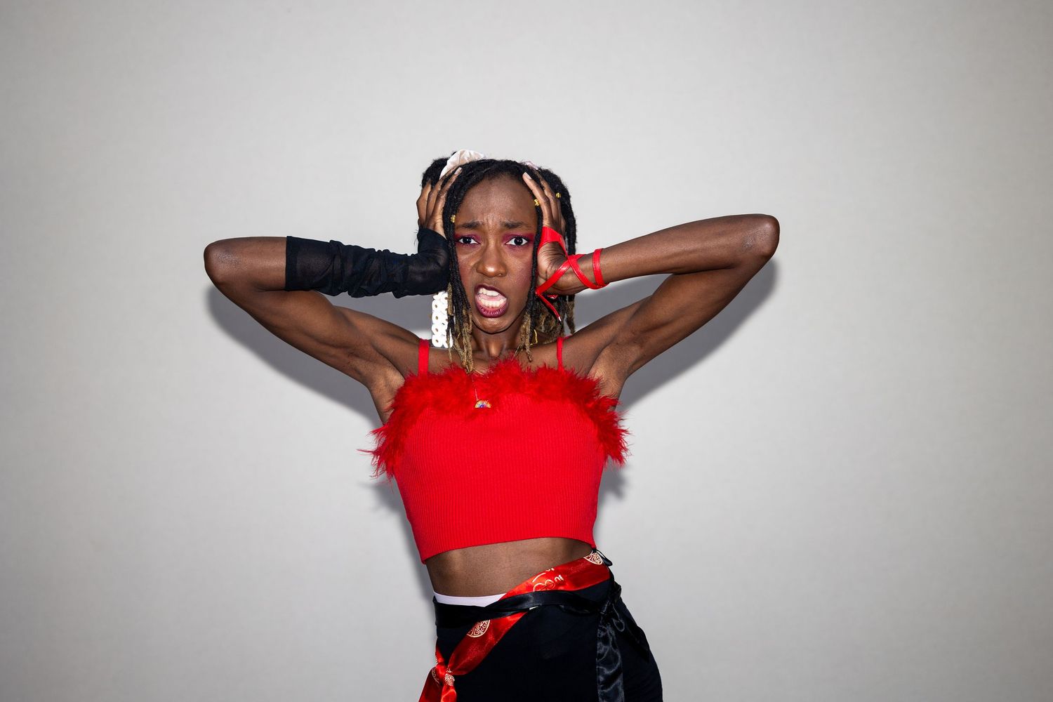 Young woman in red crop top with feathers, black shorts, and pigtails poses with hands behind head.