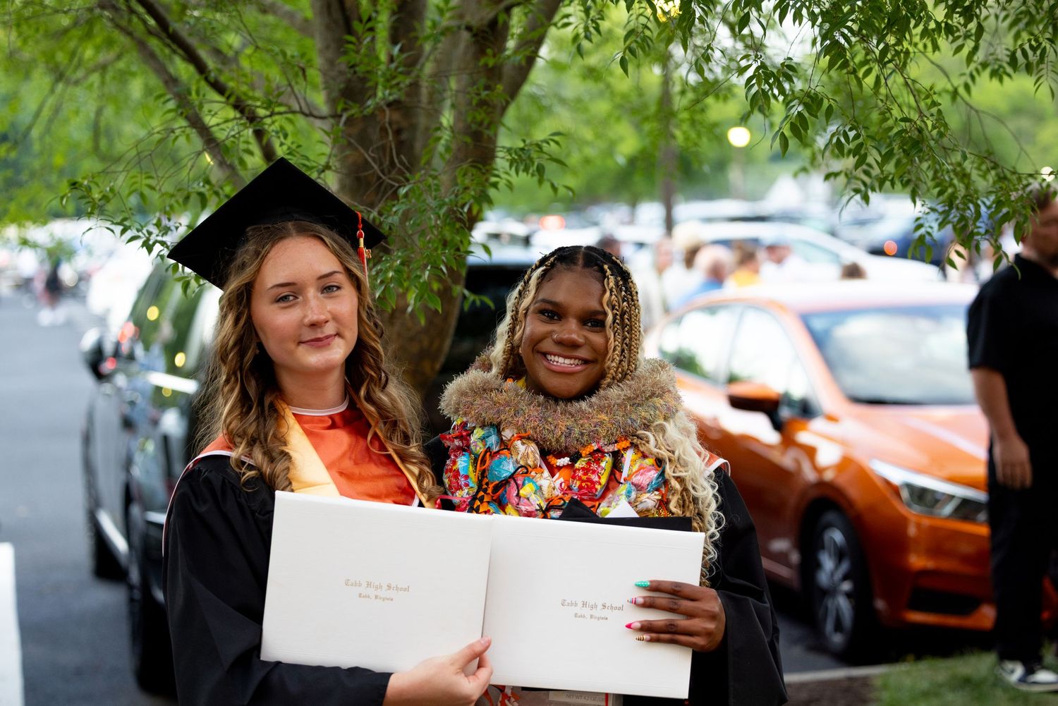 Two graduates in cap and gown smile while holding diplomas outdoors at a graduation ceremony.