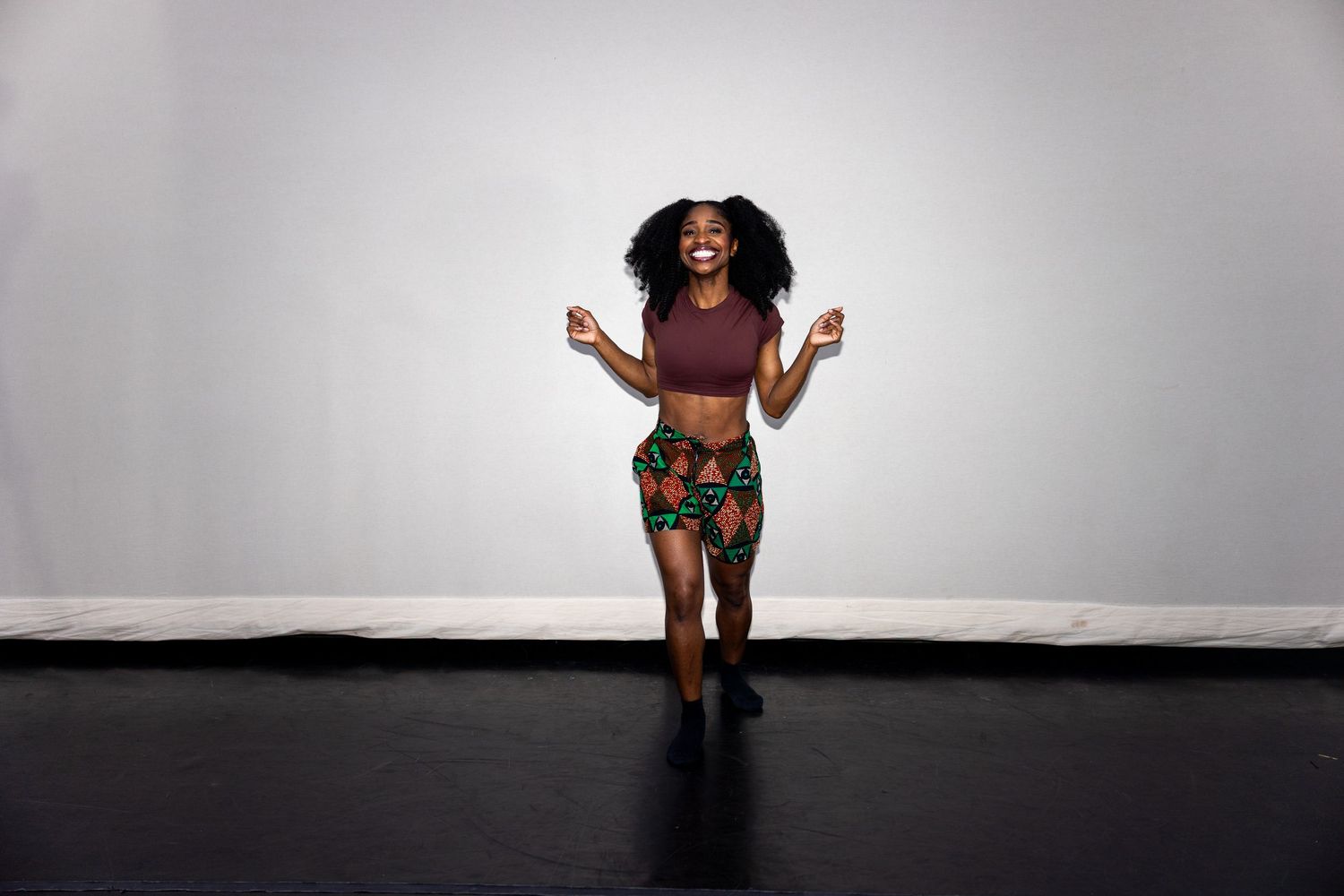 Black female dancer celebrates joyfully in maroon crop top and vibrant African print shorts.