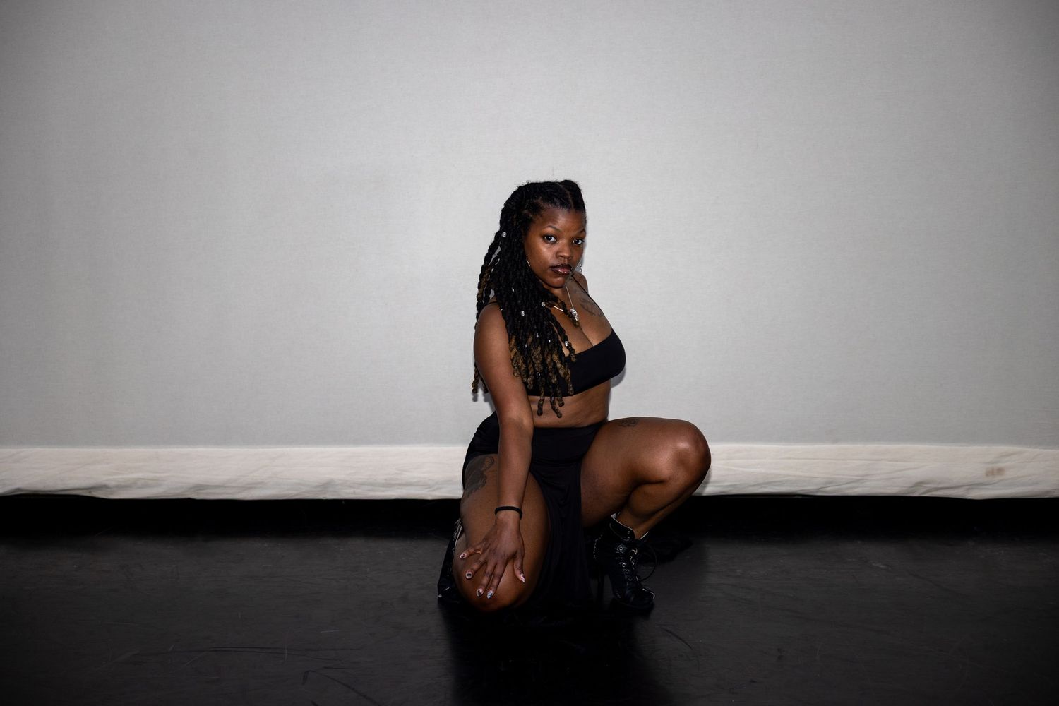 A dancer in black bralette and skirt crouches gracefully on a dark studio floor.