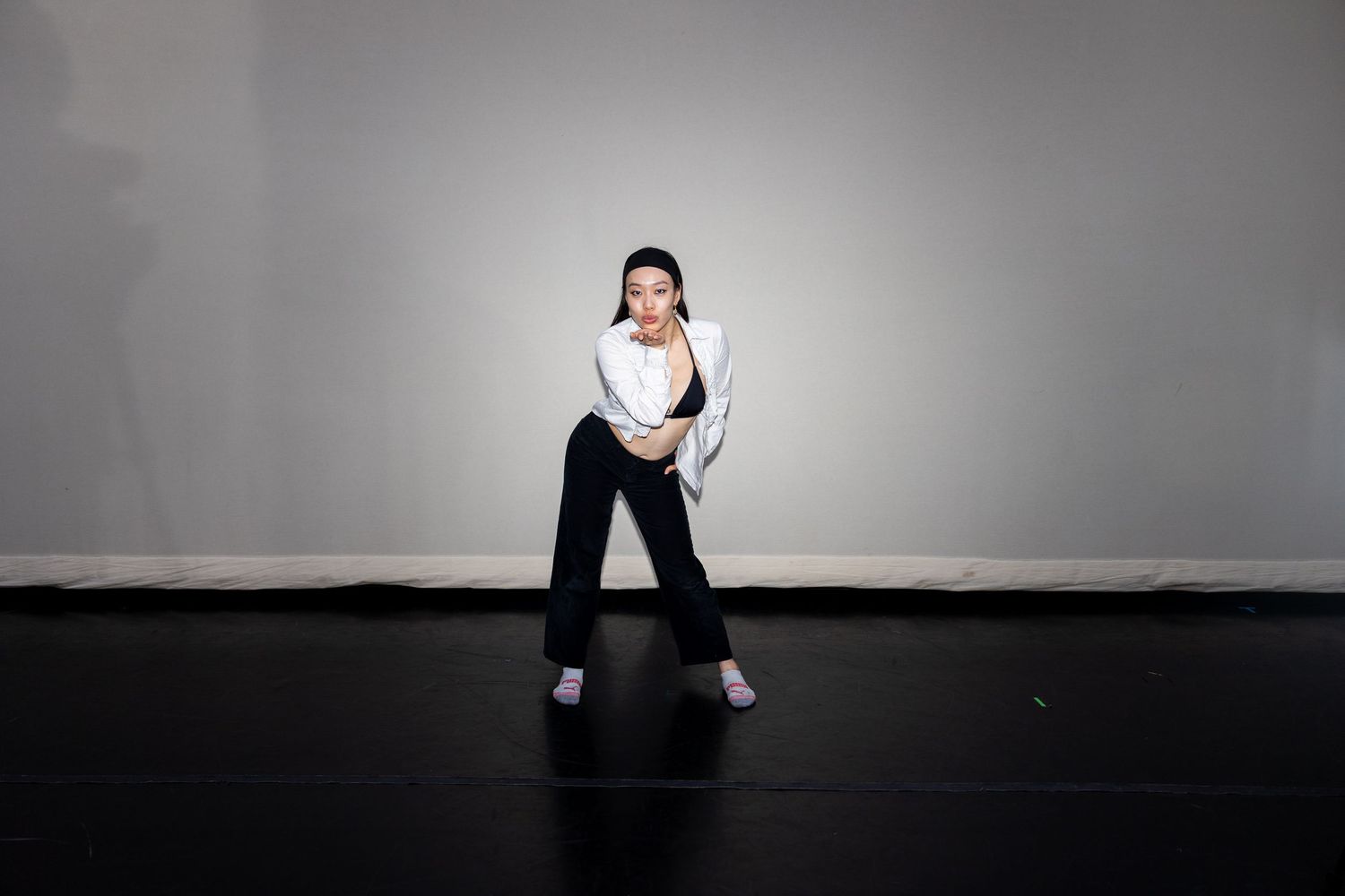 A dancer in white shirt and black pants strikes a dramatic pose on a dark stage floor against a gray wall.
