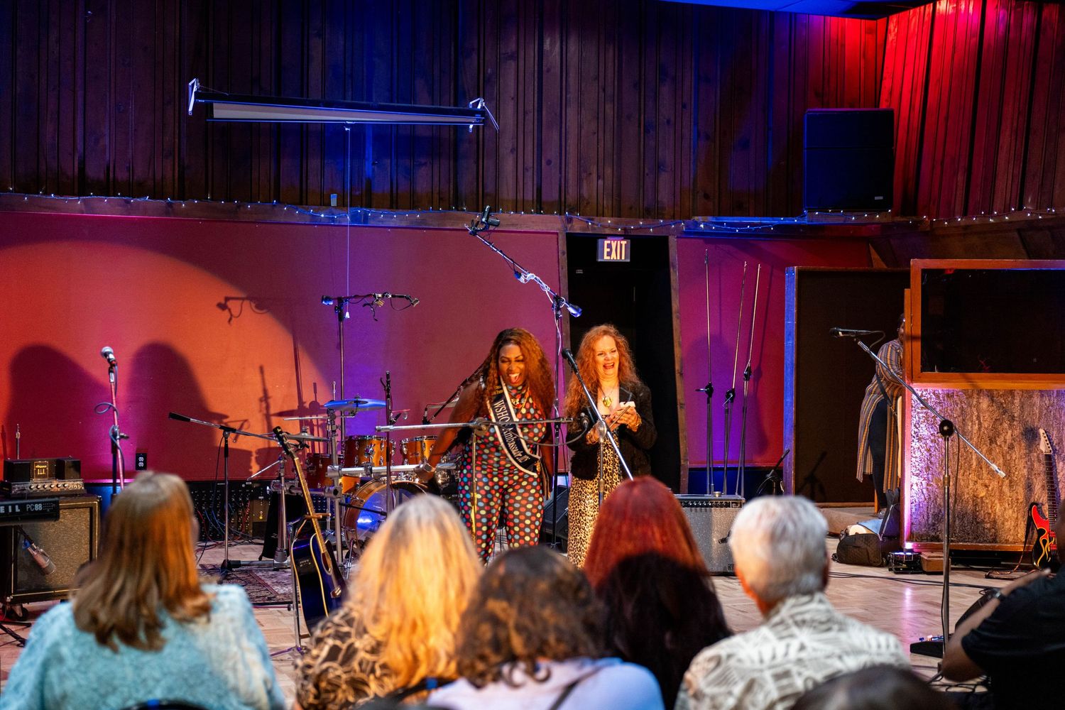 Two female performers on stage at an intimate venue with red lighting and audience seated in foreground.