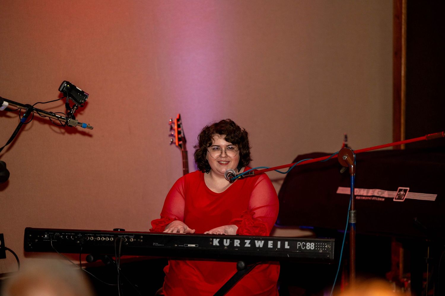 Woman in red dress smiling while playing a Kurzweil keyboard on stage with guitar in background.
