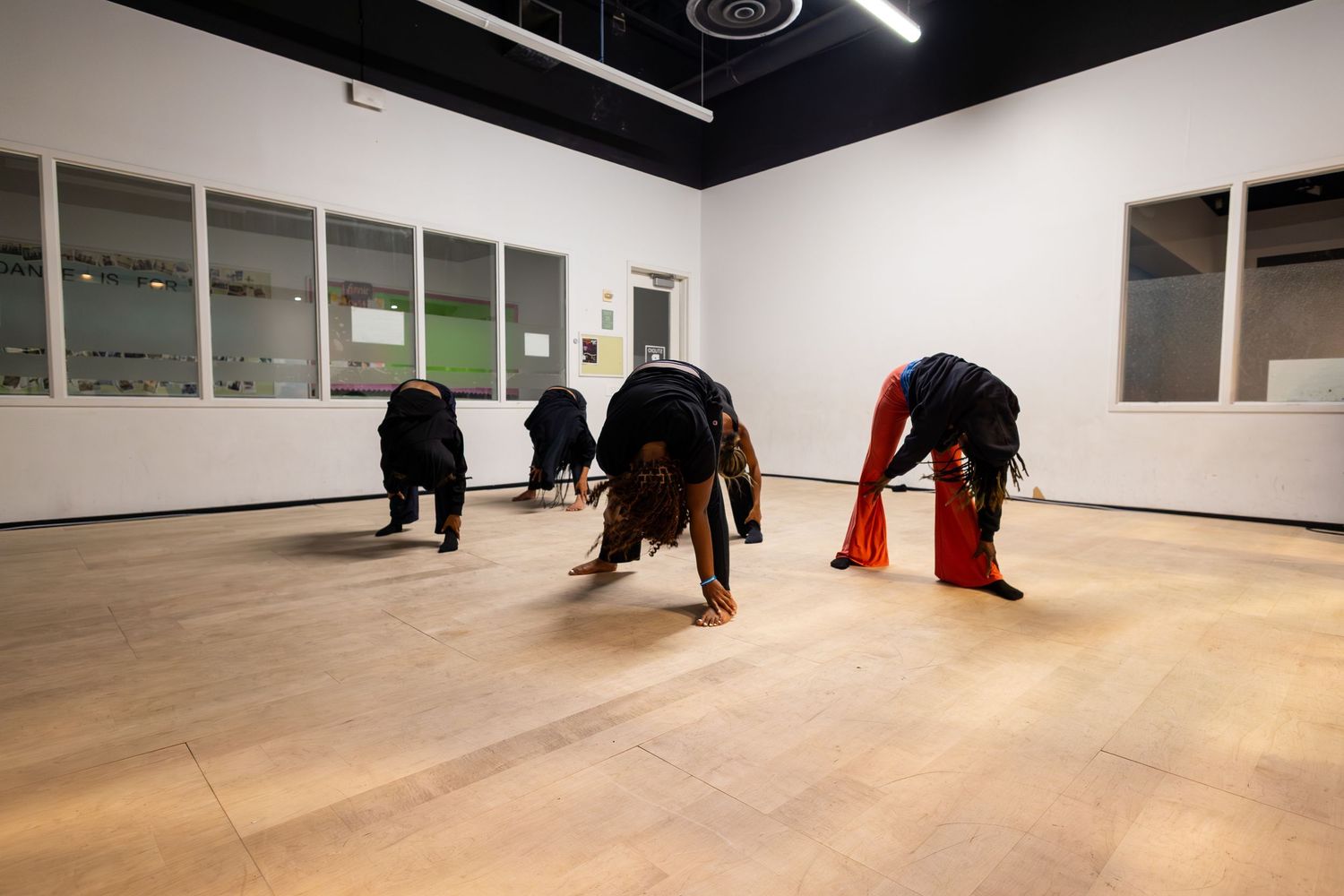 Performers in black and red costumes crouch low during a contemporary dance in a white studio.