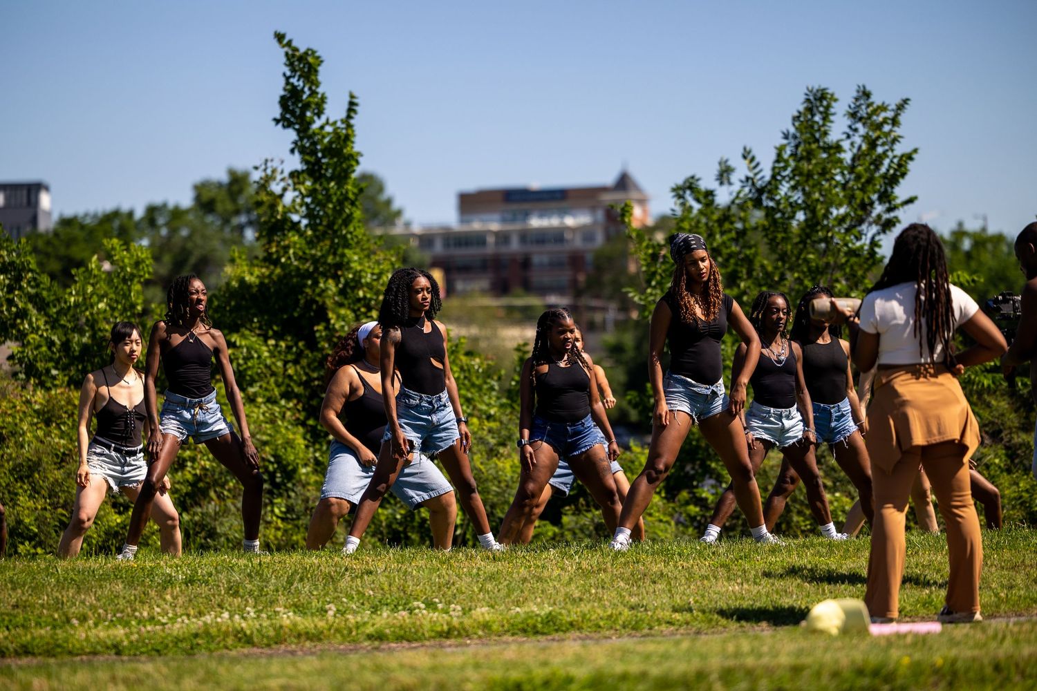 A dance instructor in a tan outfit leads a group of young women in matching black tops through a choreographed routine.