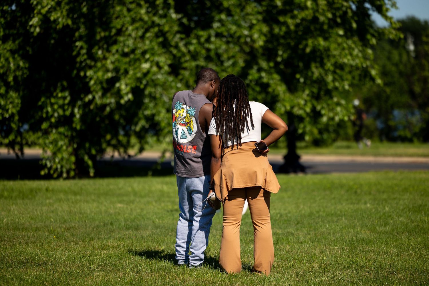 Couple standing together on green grass in a sunny park, woman in tan outfit and man in gray shirt.