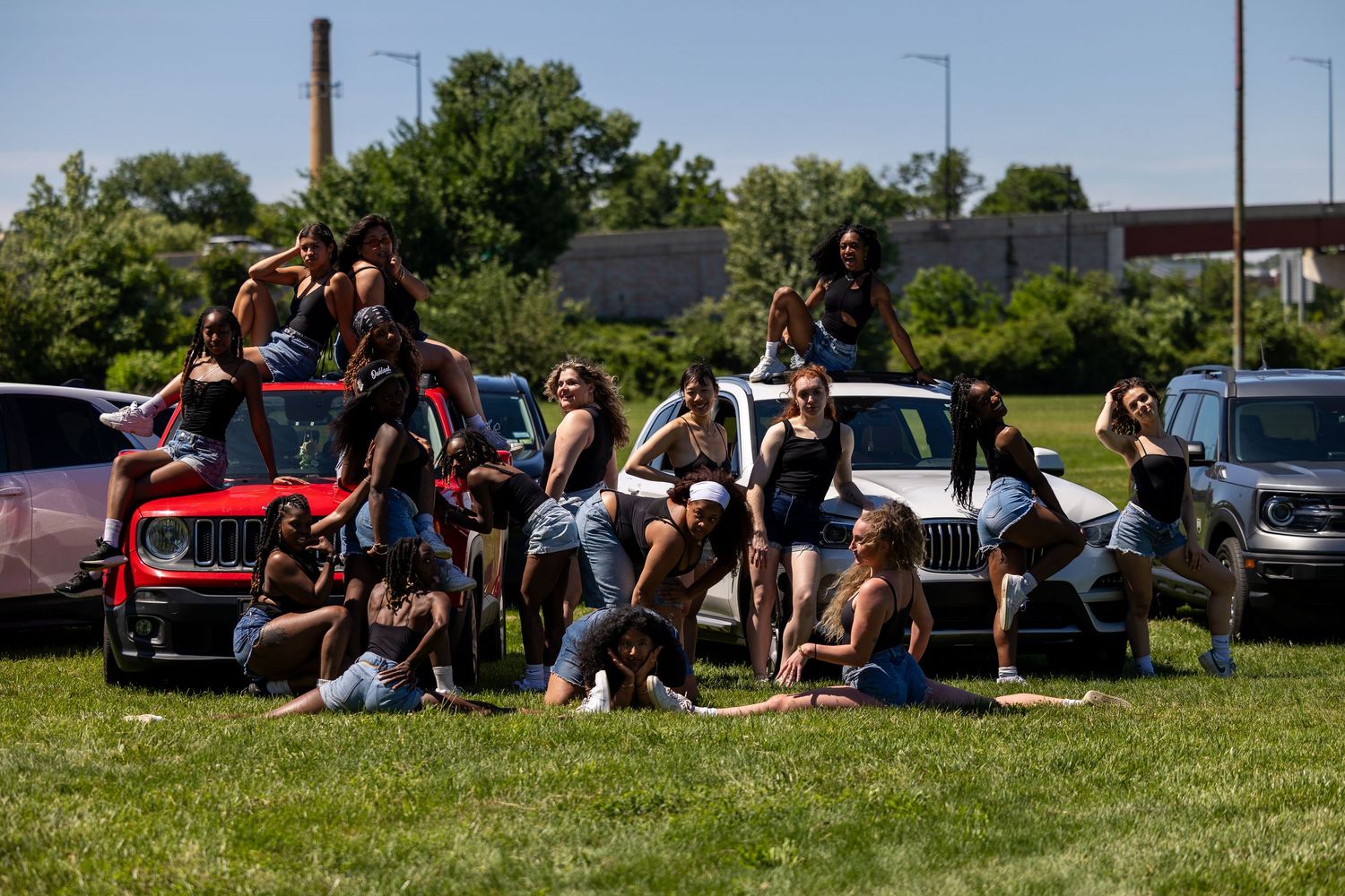 Group of young women posing together on grass near parked cars on a sunny day.