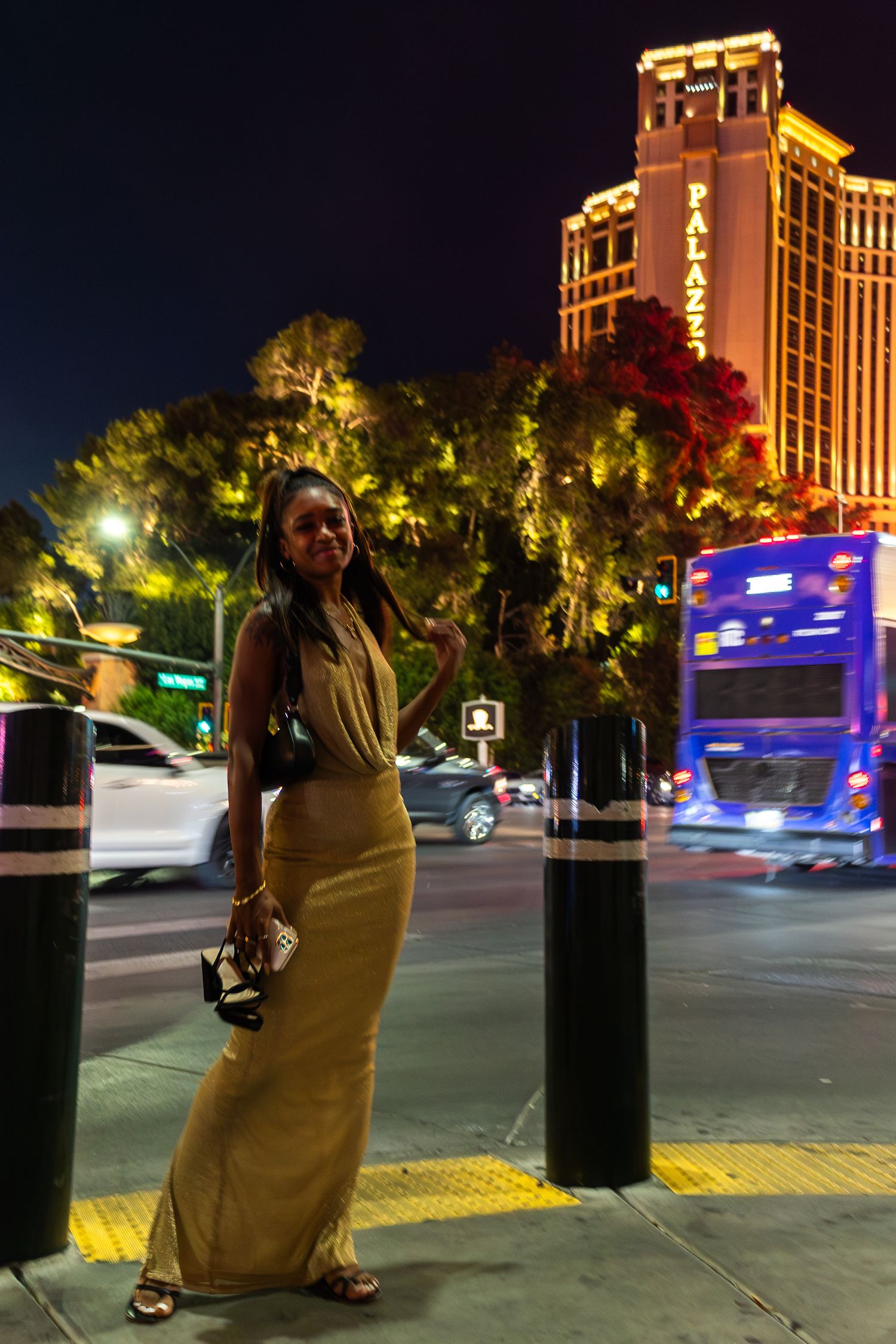 Illuminated city buildings and palm trees at night with motion blur from passing vehicles.