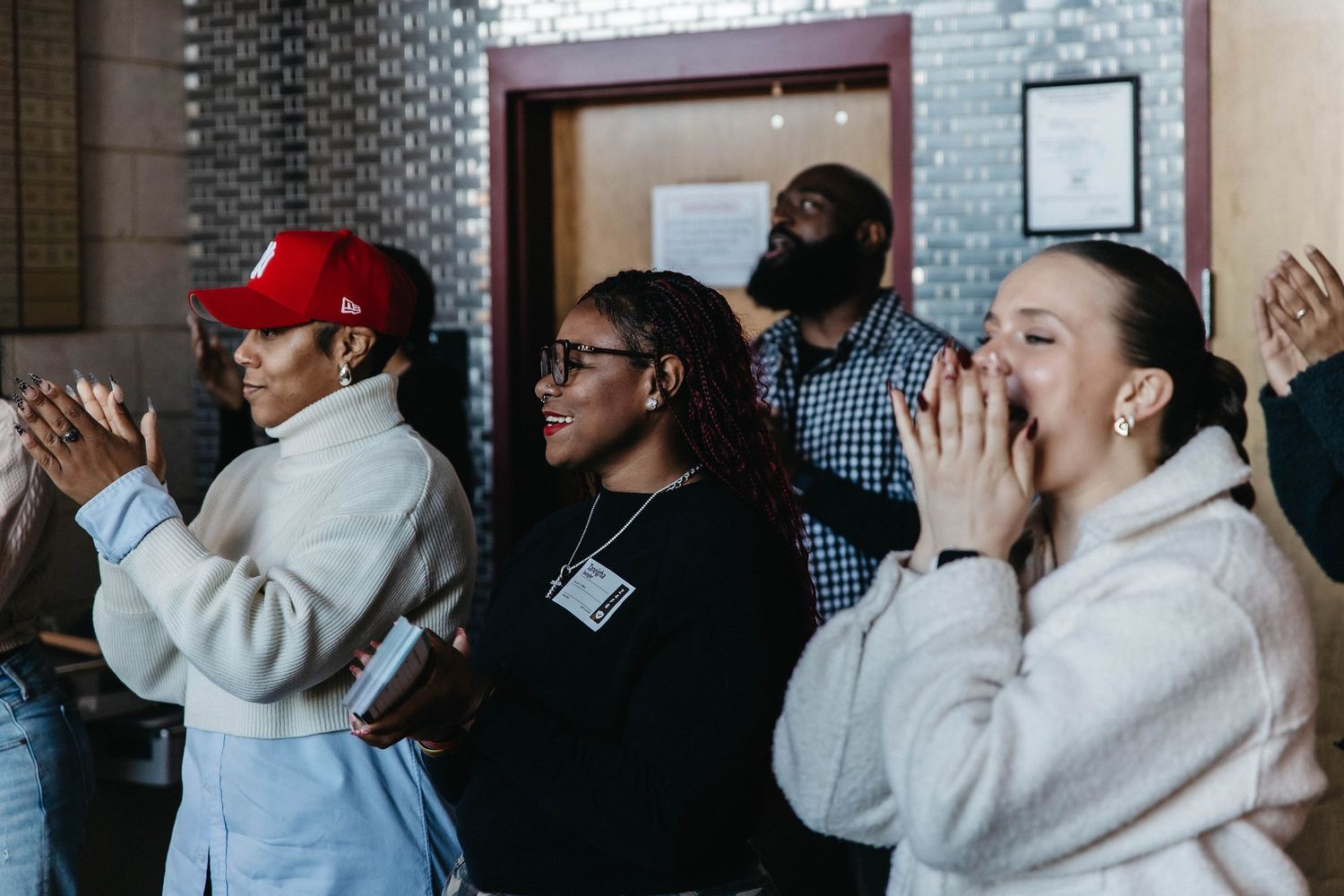 A diverse group of people stand together clapping and celebrating in an indoor setting.