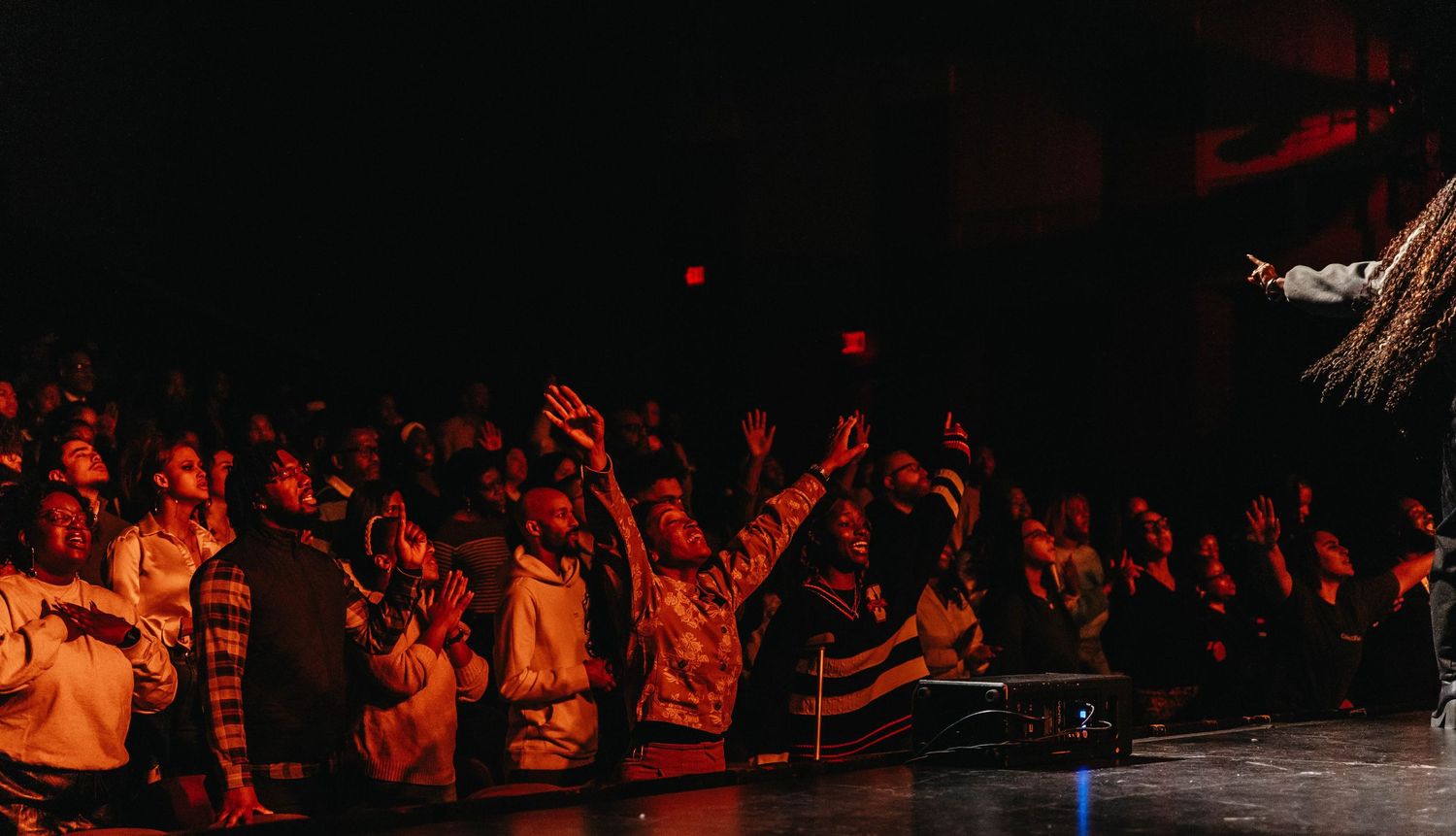 Crowd of concert-goers with raised arms illuminated by red lighting at a live music performance.