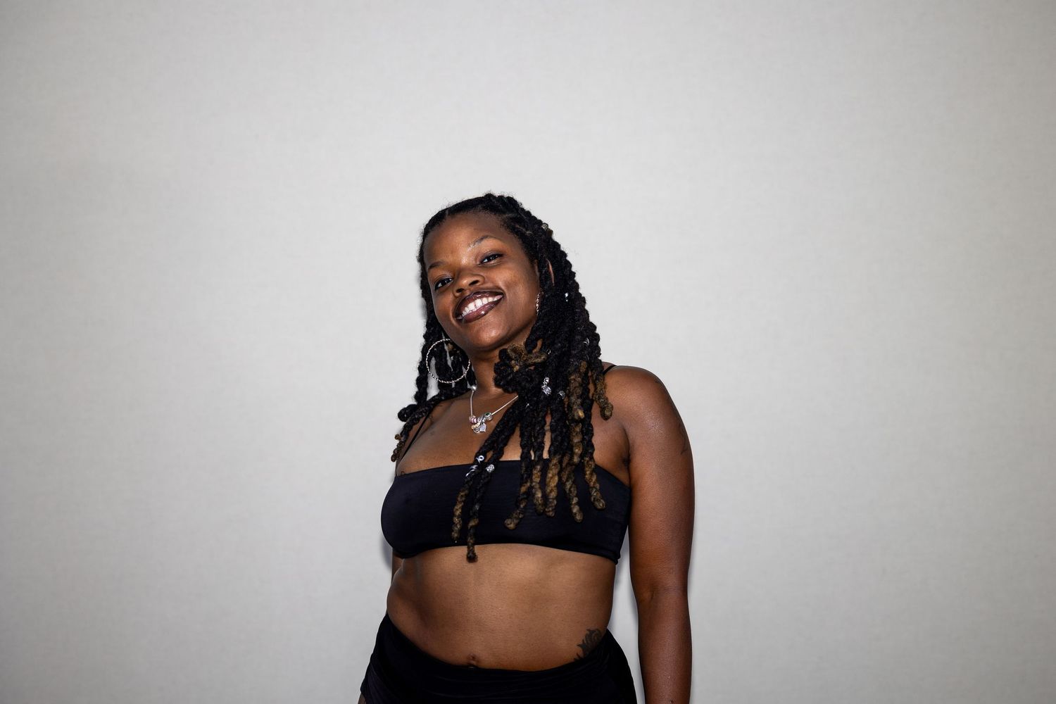 Smiling young woman with braided hair wearing a black crop top and pants against a white background.