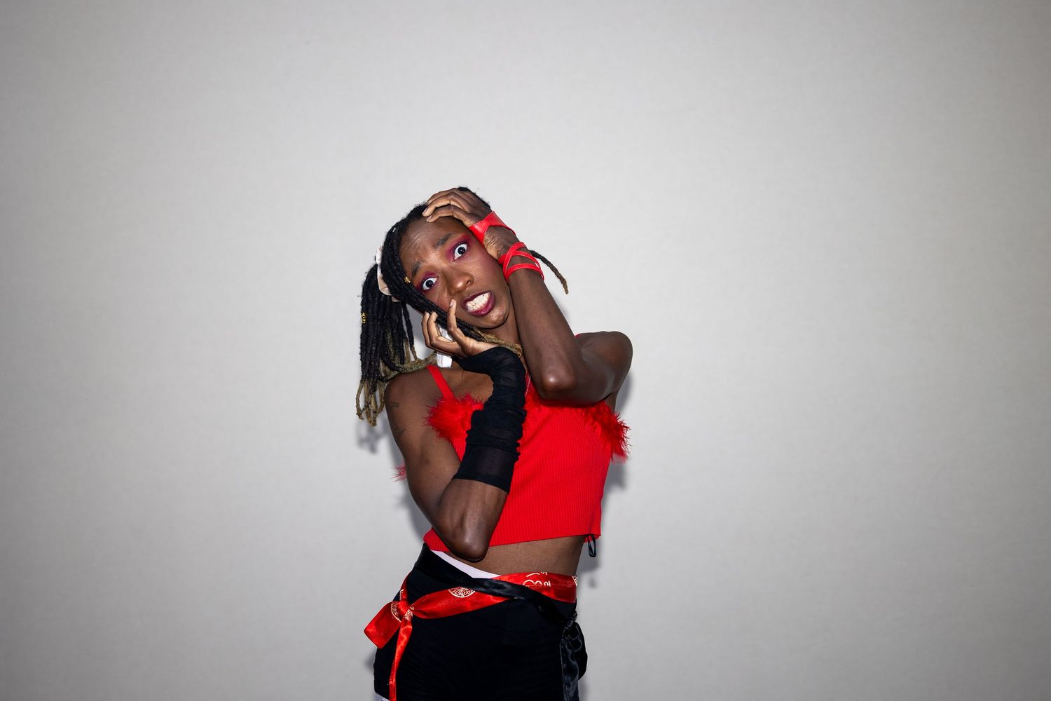 Expressive performer in red crop top and black fingerless gloves striking a playful pose against gray background.