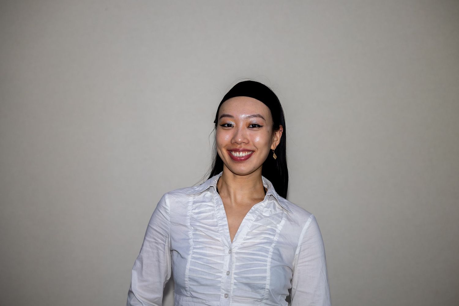 Smiling Asian woman with dark hair wearing a white ruffled button-up shirt poses against a wall.