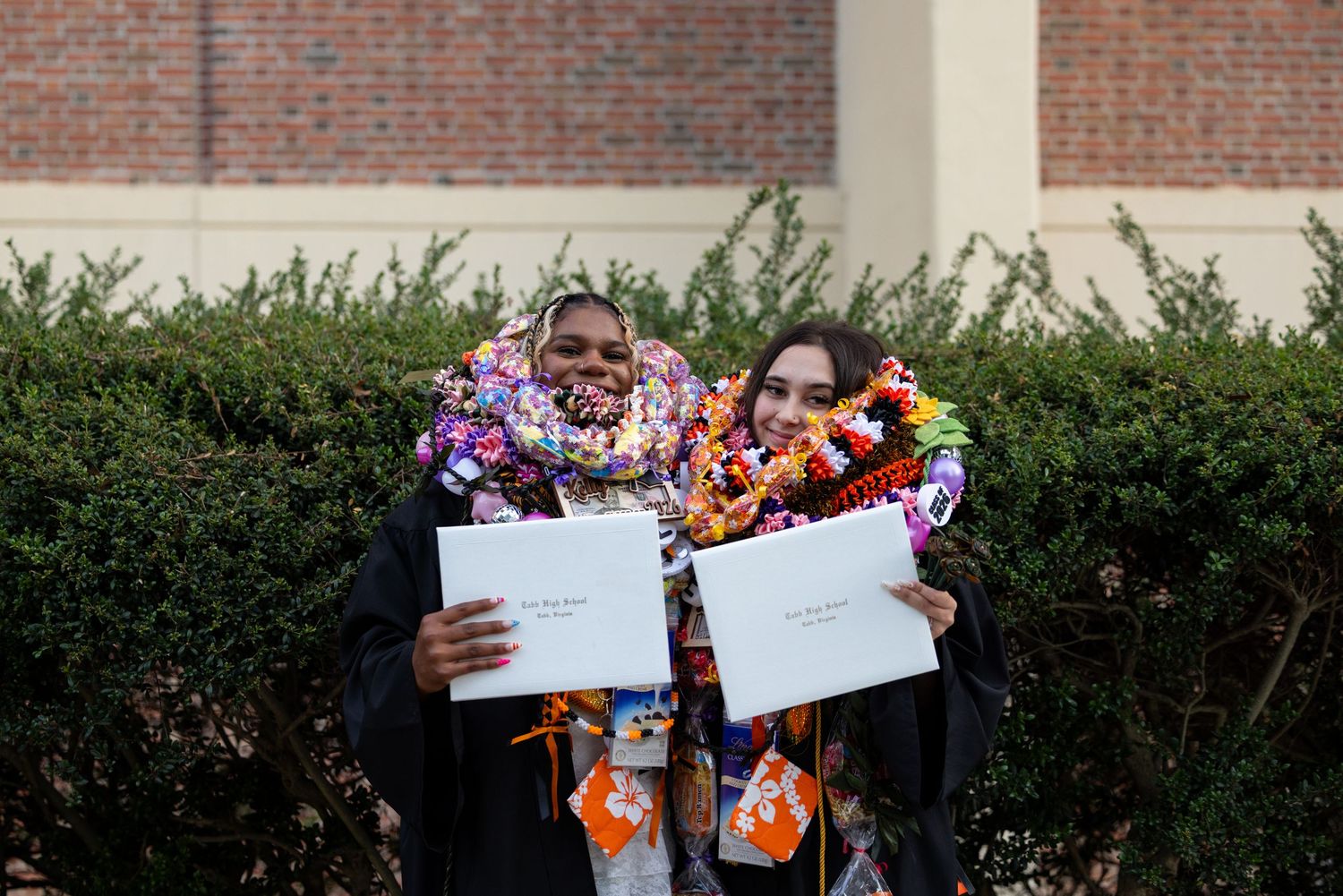 Two graduates in black gowns and floral leis proudly hold diplomas while posing together outdoors.