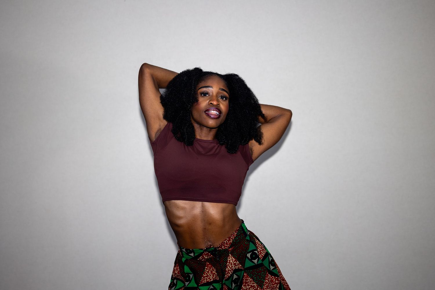 A fit woman in a burgundy crop top and floral skirt poses with arms raised against a gray background.