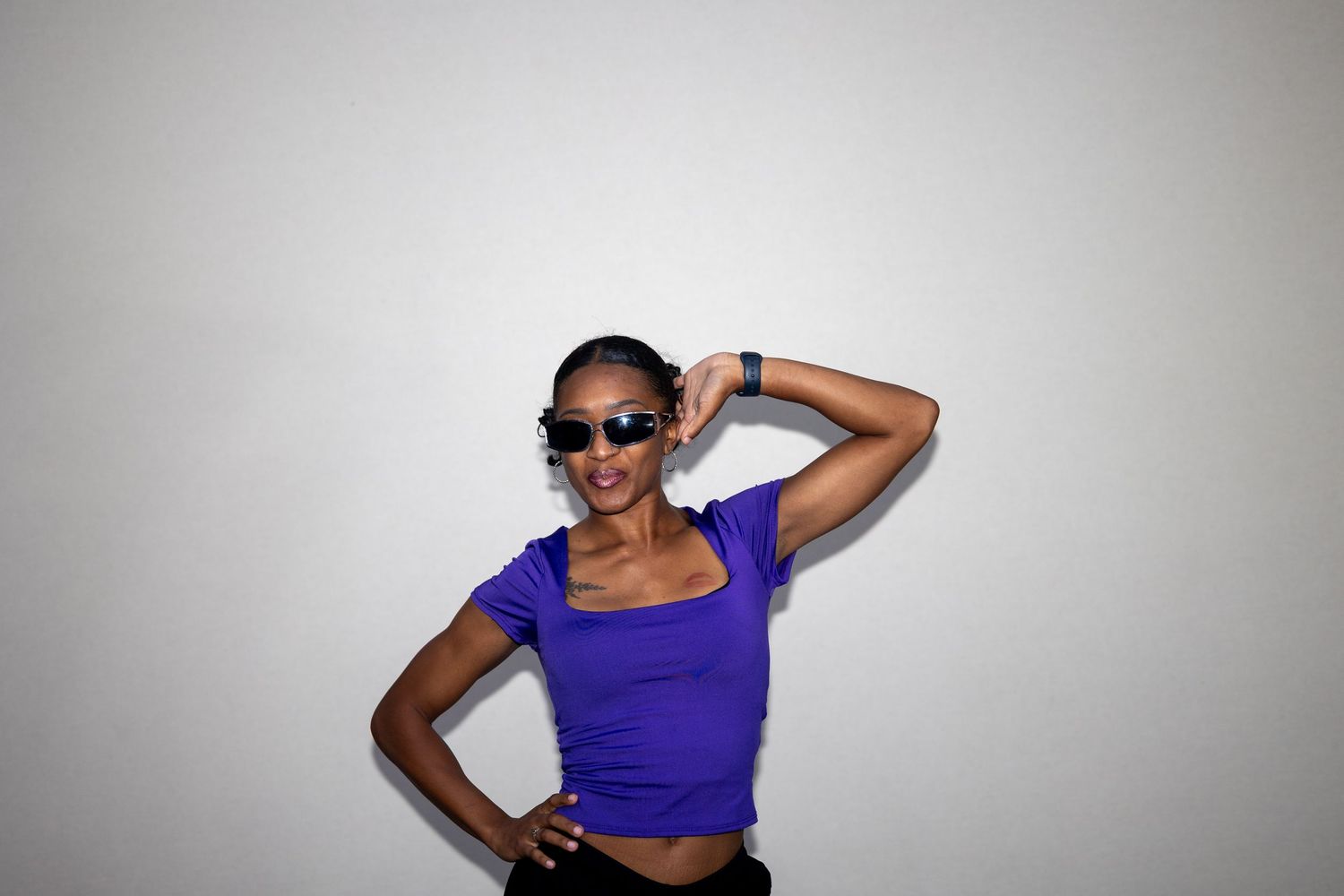 A fashionable woman in a purple crop top and sunglasses strikes a fun pose against a white background.