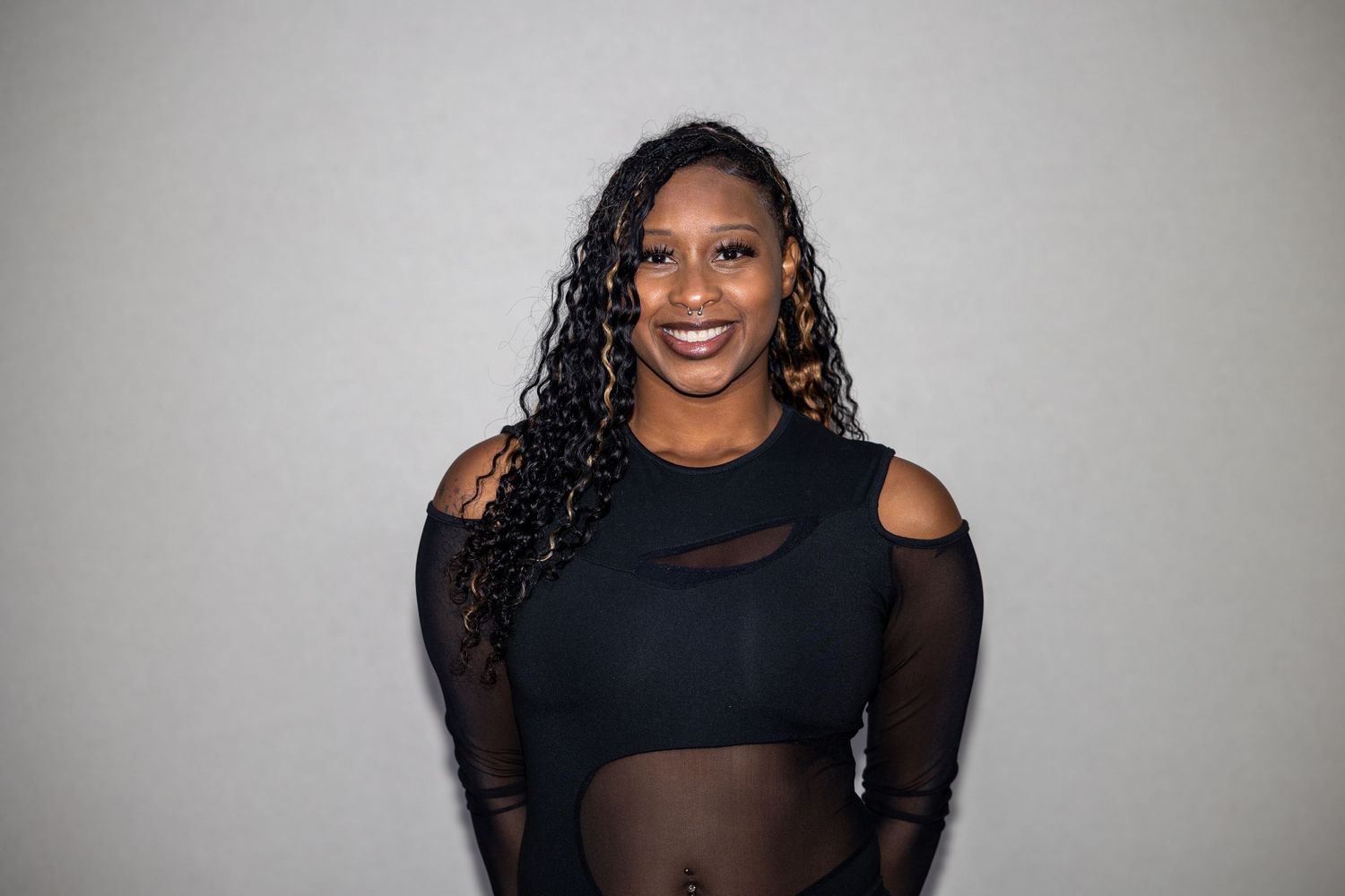 Smiling Black woman with long curly hair wearing a black mesh crop top poses against a gray background.