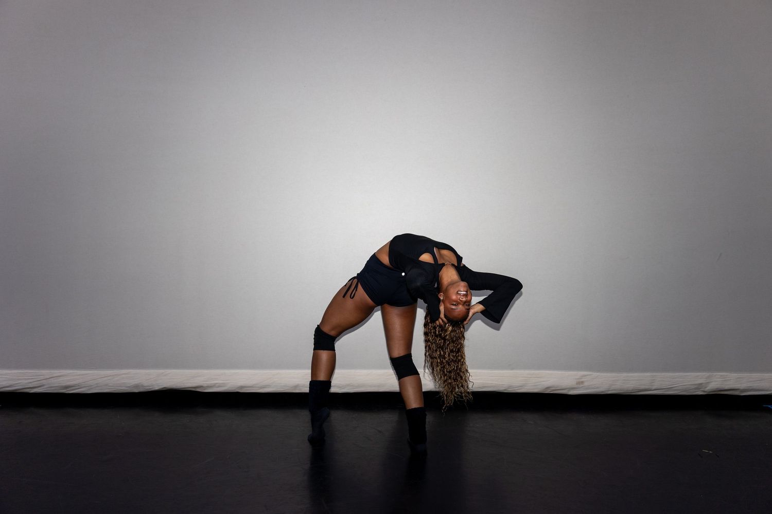 A dancer in a black outfit bends backward dramatically on a dark studio floor.