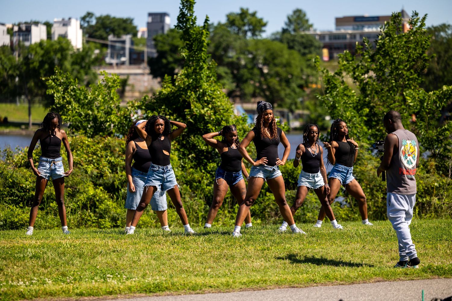 A dance instructor watches as a group of young women rehearse a synchronized routine in an outdoor park setting.