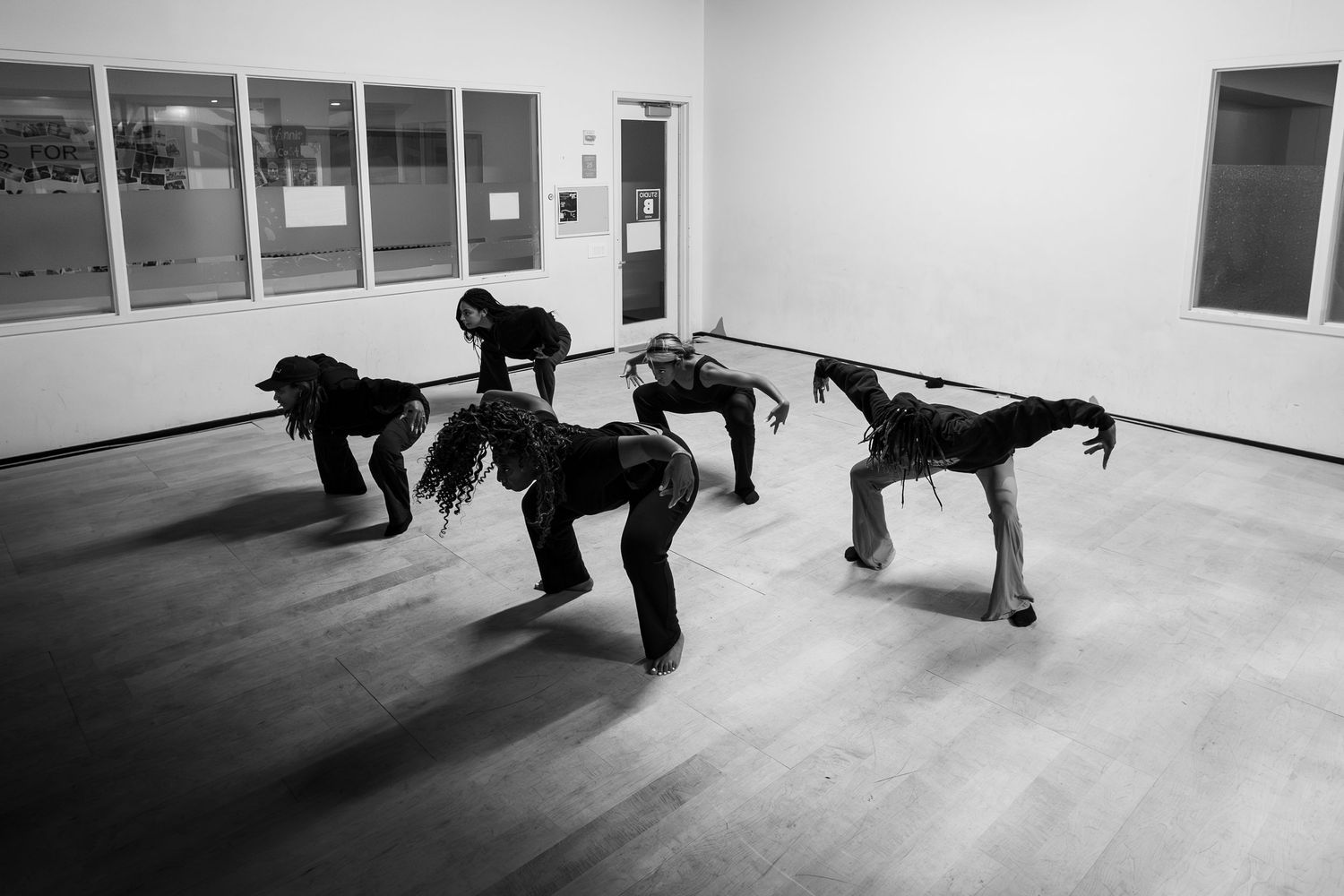 Black and white photo of four dancers bent forward in unison wearing all black in a dance studio.