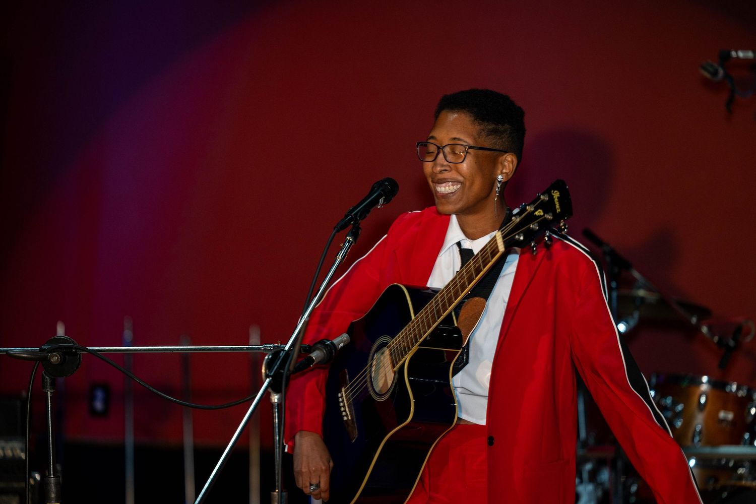 Young musician in red blazer smiles while playing acoustic guitar on stage at a live performance.