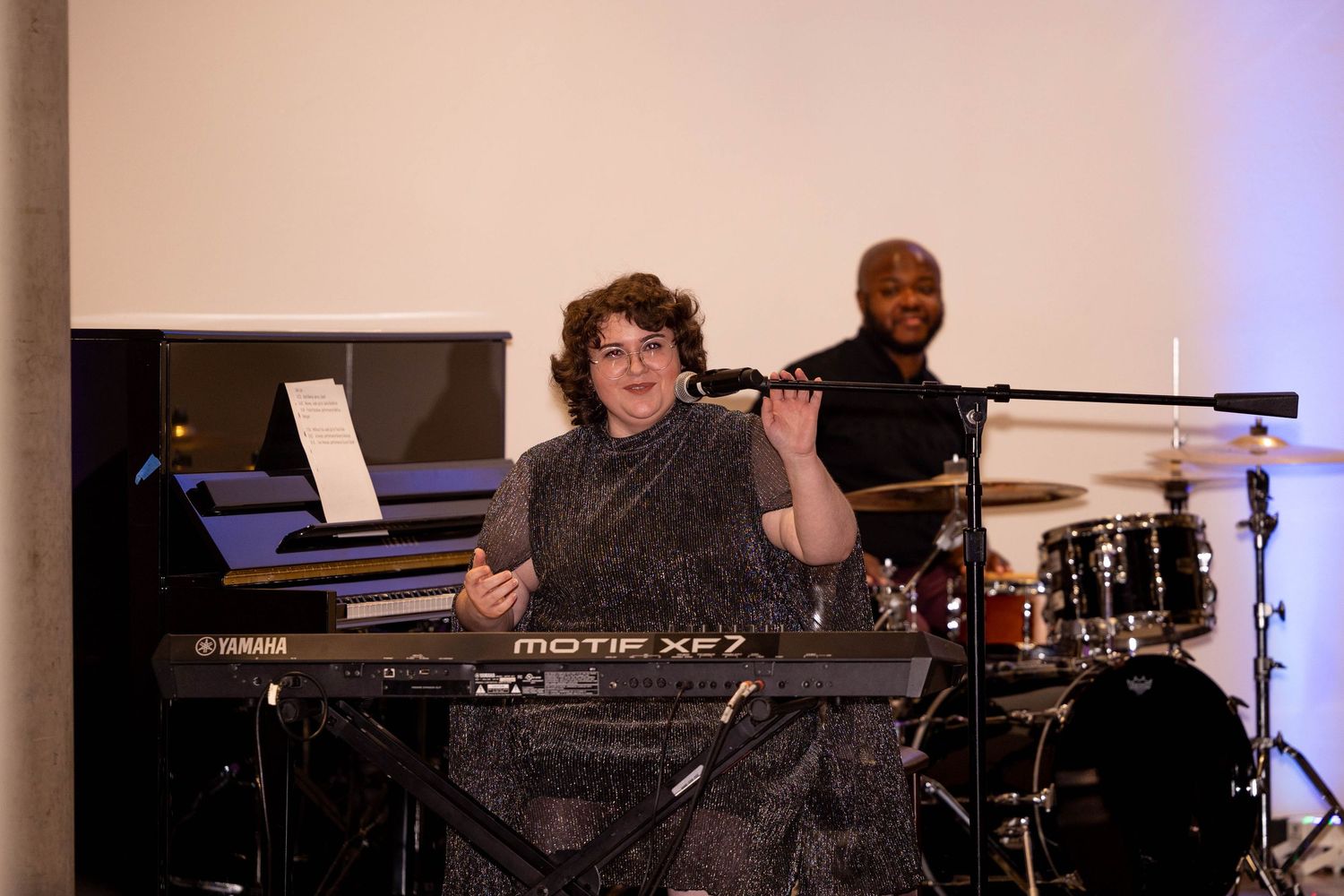 Female musician smiling at Yamaha Motif XF7 keyboard with drummer performing in background.