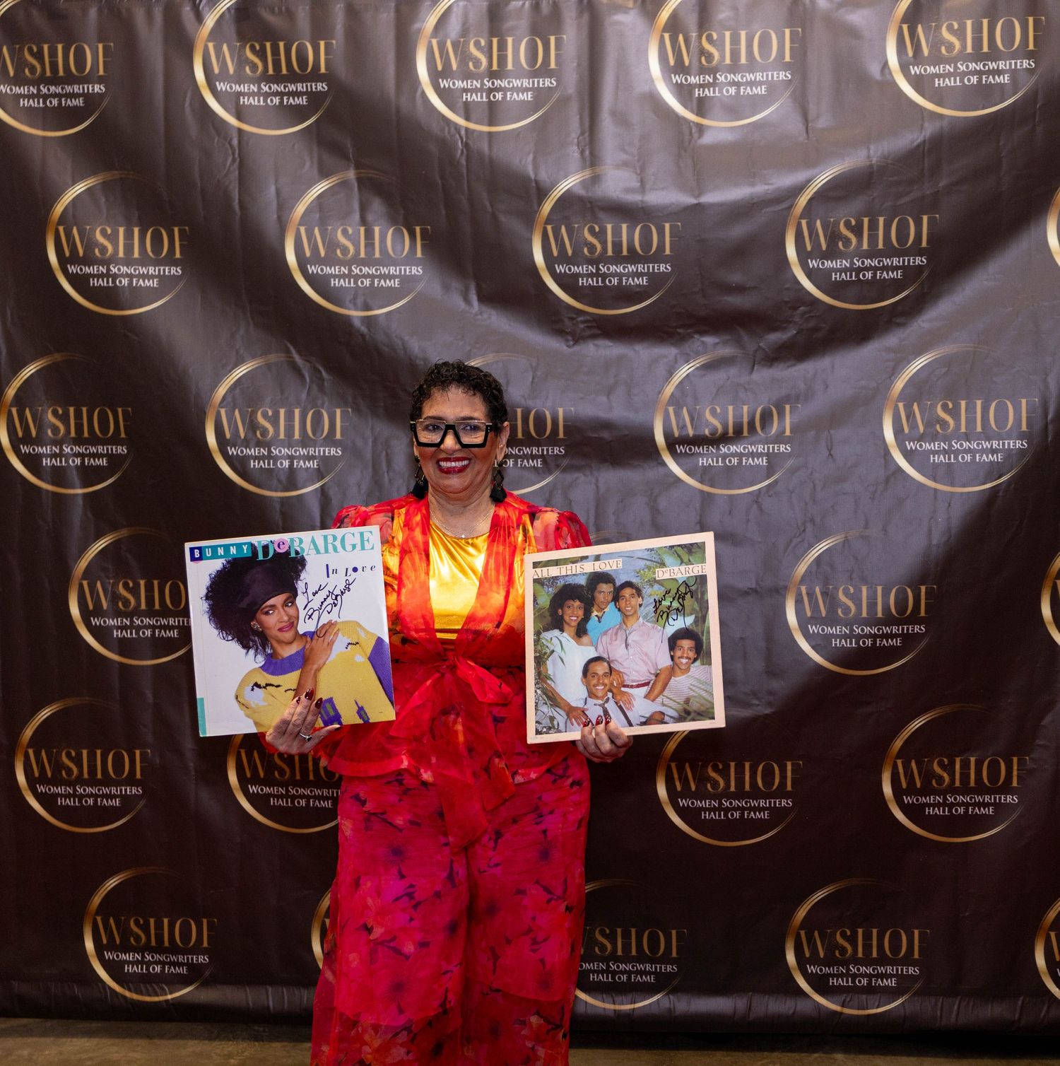 Woman in red outfit holds vinyl records at WSHOF Women Songwriters Hall of Fame event backdrop.