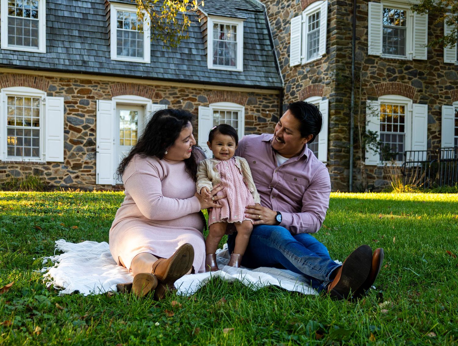 Young family with baby girl sitting on blanket on lawn in front of historic stone home.
