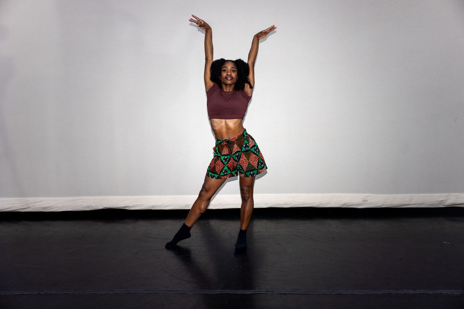 A dancer in African print skirt raises arms overhead in an expressive dance pose on stage.