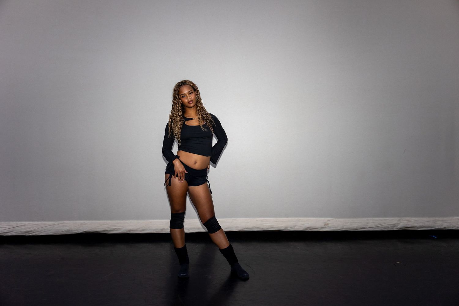 A dancer in black crop top, shorts, and knee-high boots poses confidently in a studio setting.