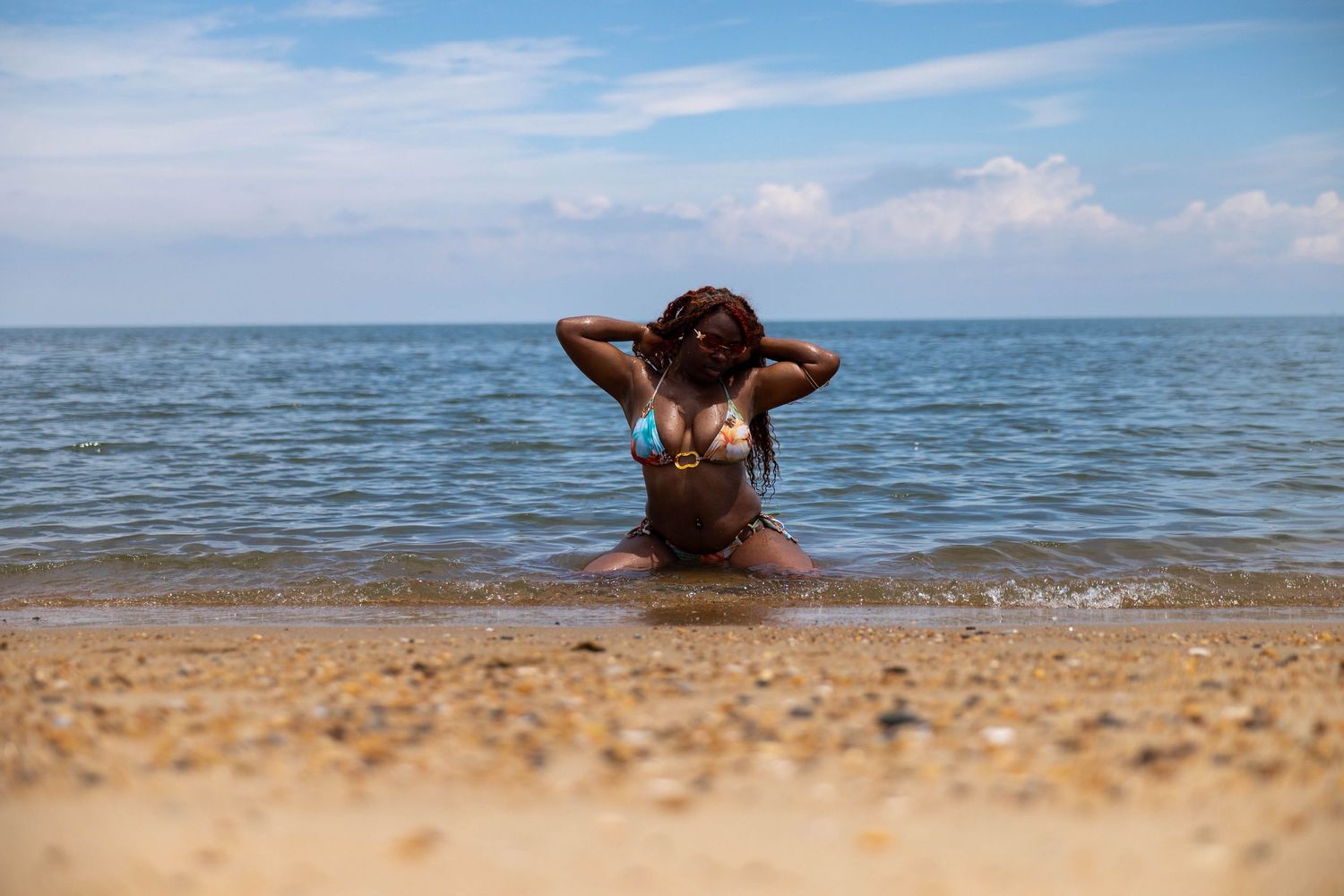 A woman in a bikini sits in shallow ocean waves on a sandy beach, facing the sea with hands behind her head.