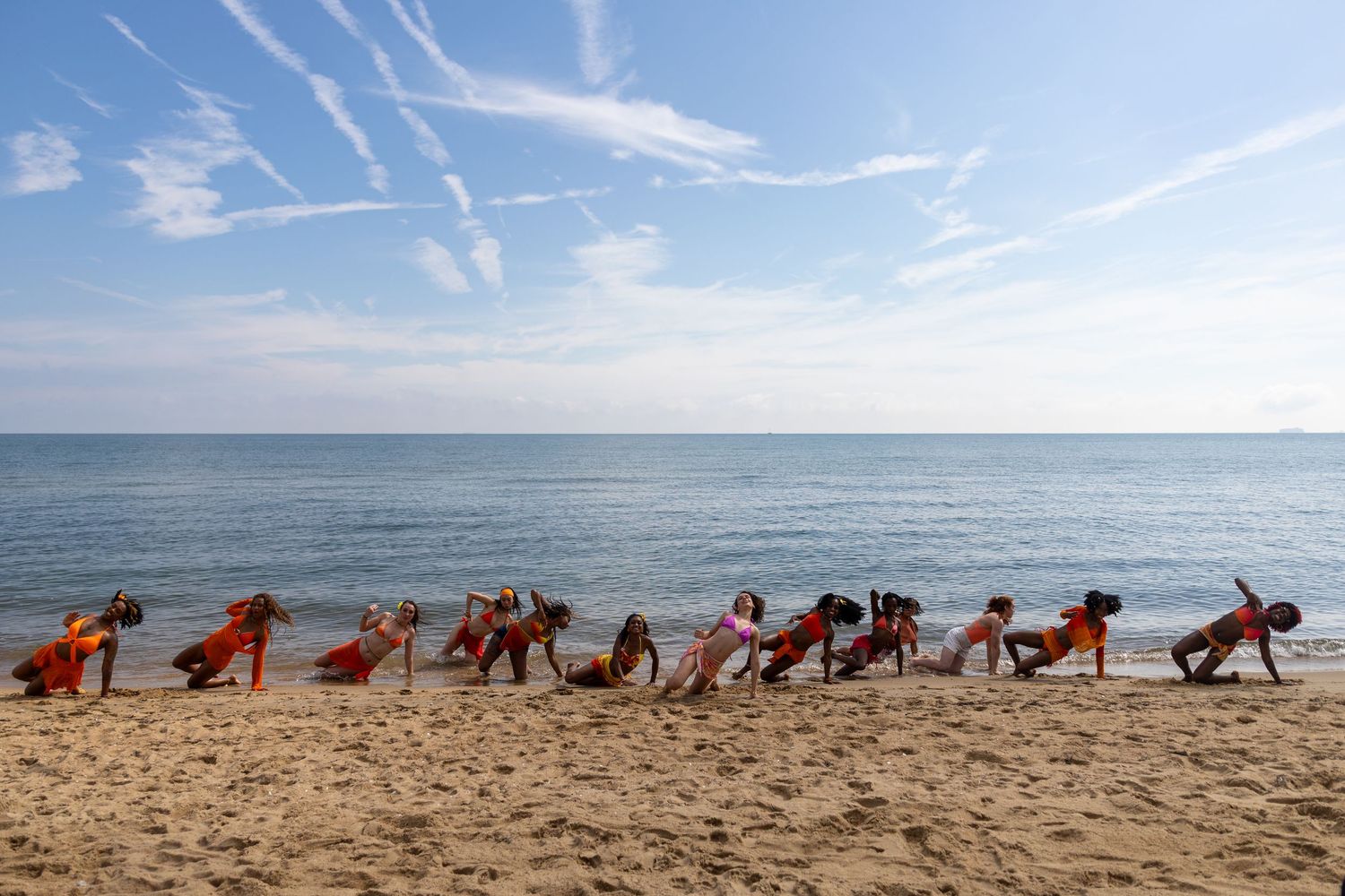 Group of people doing yoga poses in a line on a sandy beach with calm ocean and blue sky backdrop.