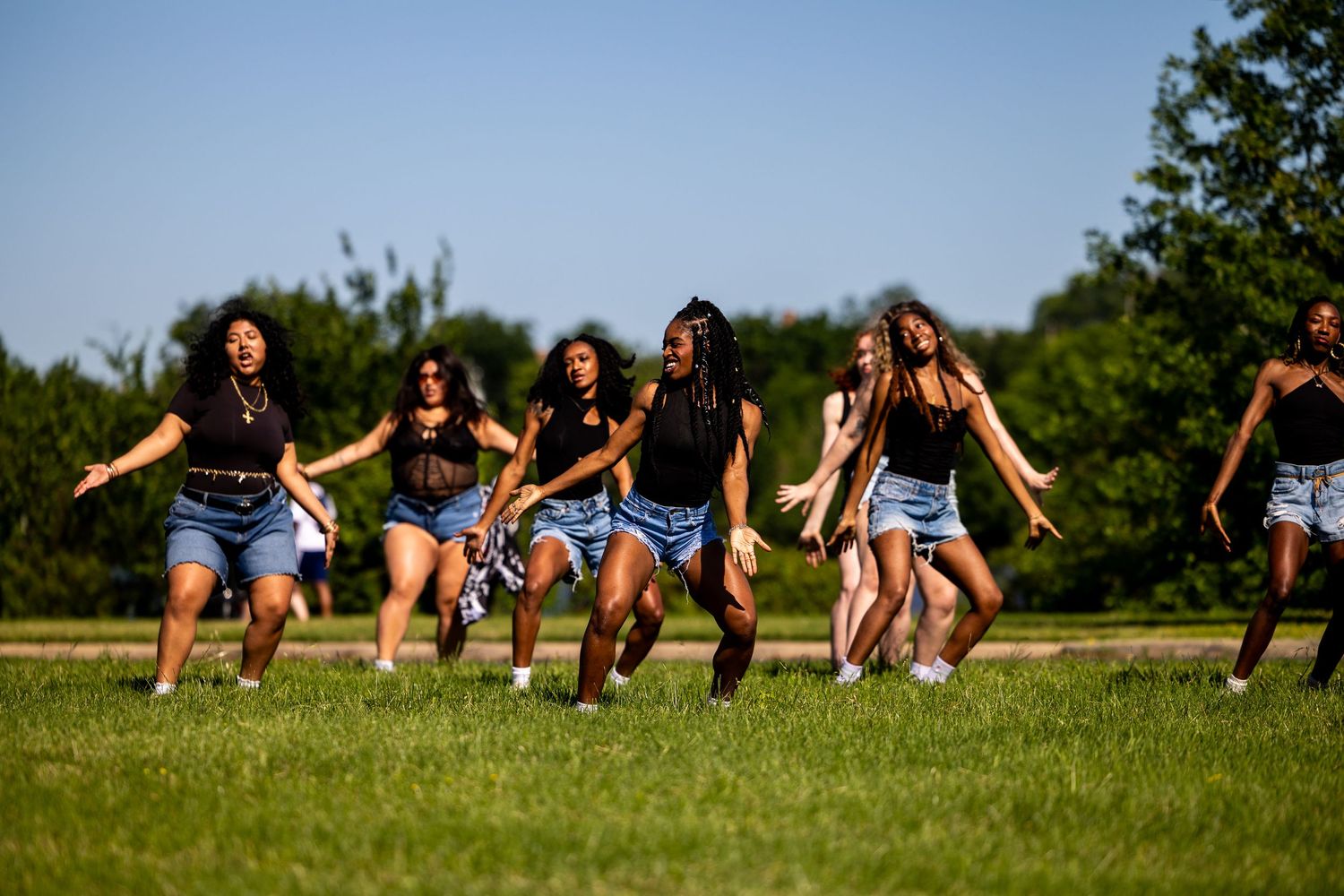 A group of young women in black tops and denim shorts dancing energetically together on a green grass field outdoors.