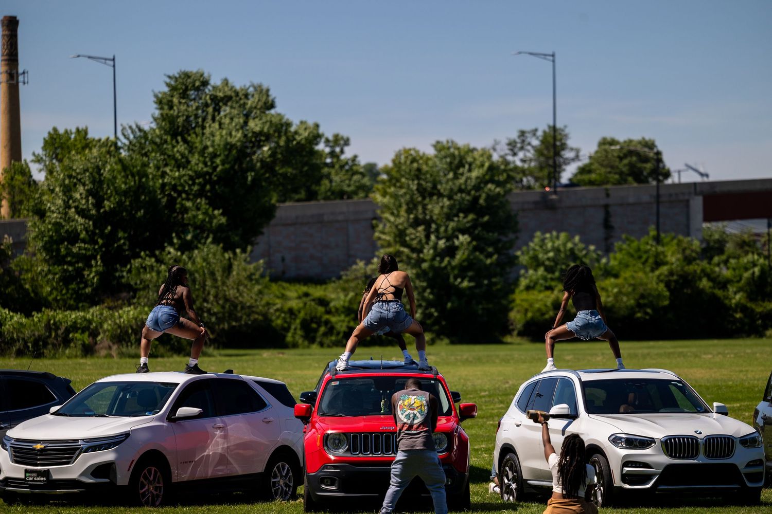 Dancers perform on car hoods at an outdoor drive-in event surrounded by trees and parked vehicles.