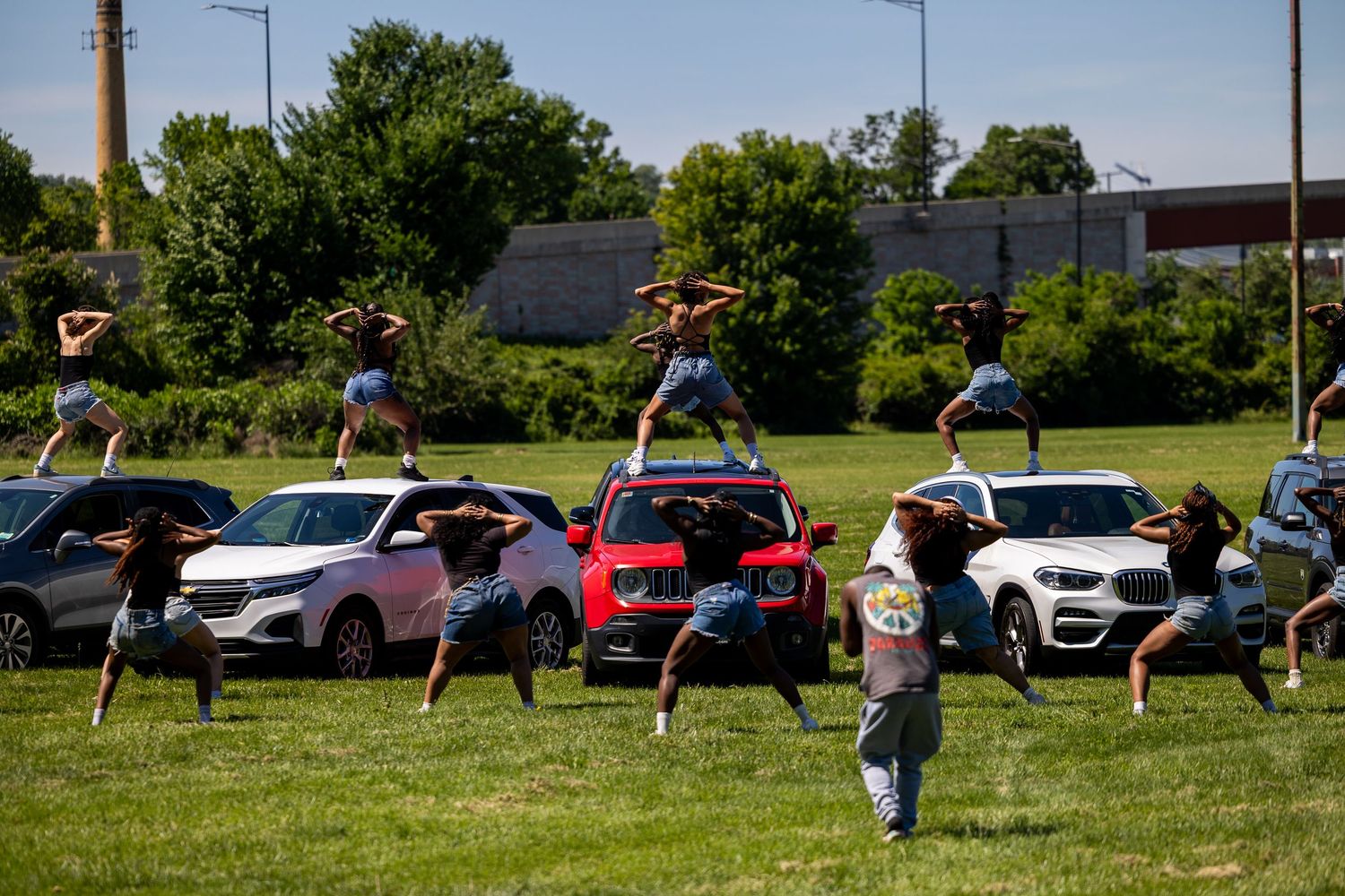 Dancers perform acrobatic stunts on cars during an outdoor event on a green field with trees in the background.