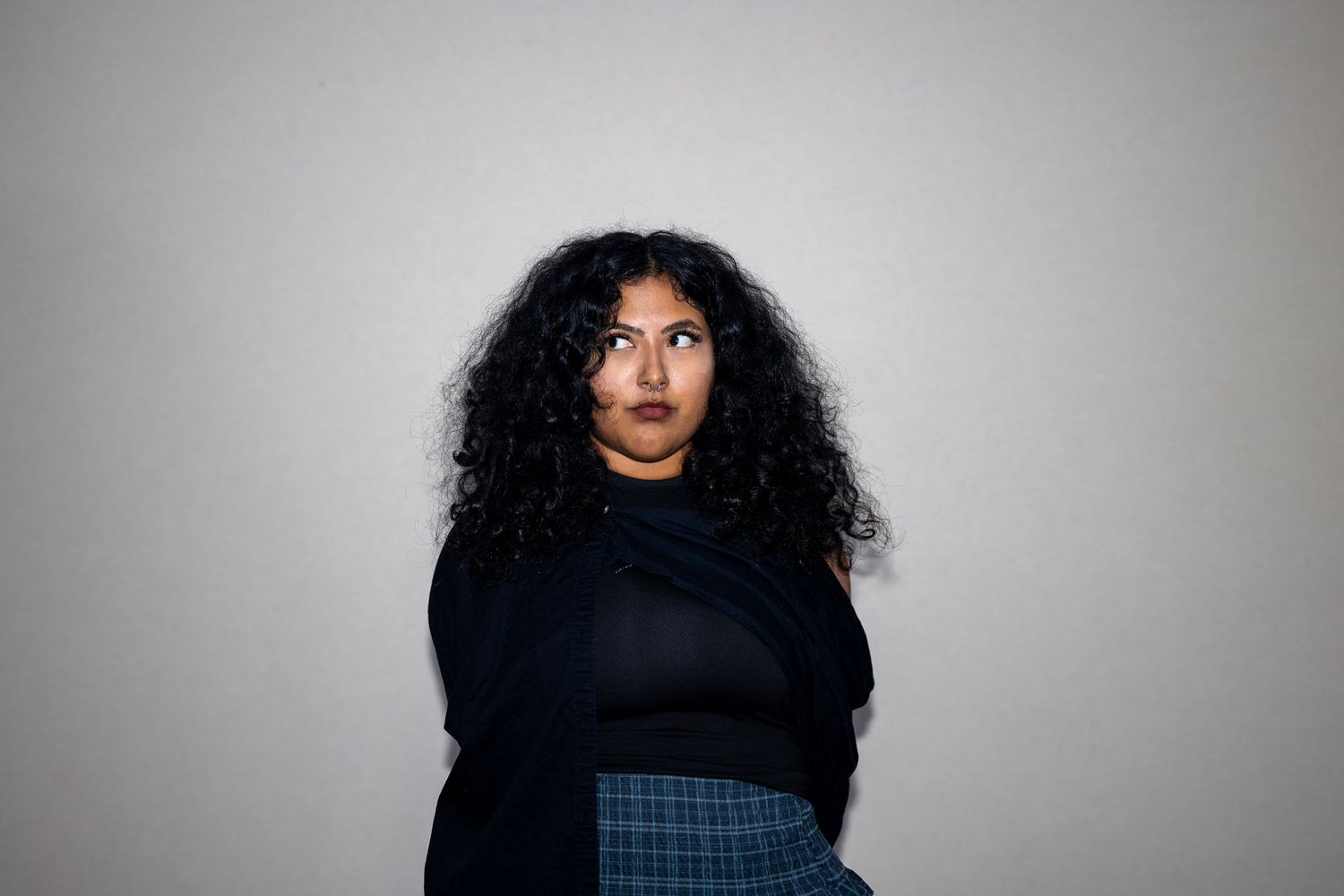 Woman with voluminous curly hair in a black top and plaid skirt poses with a serious expression.