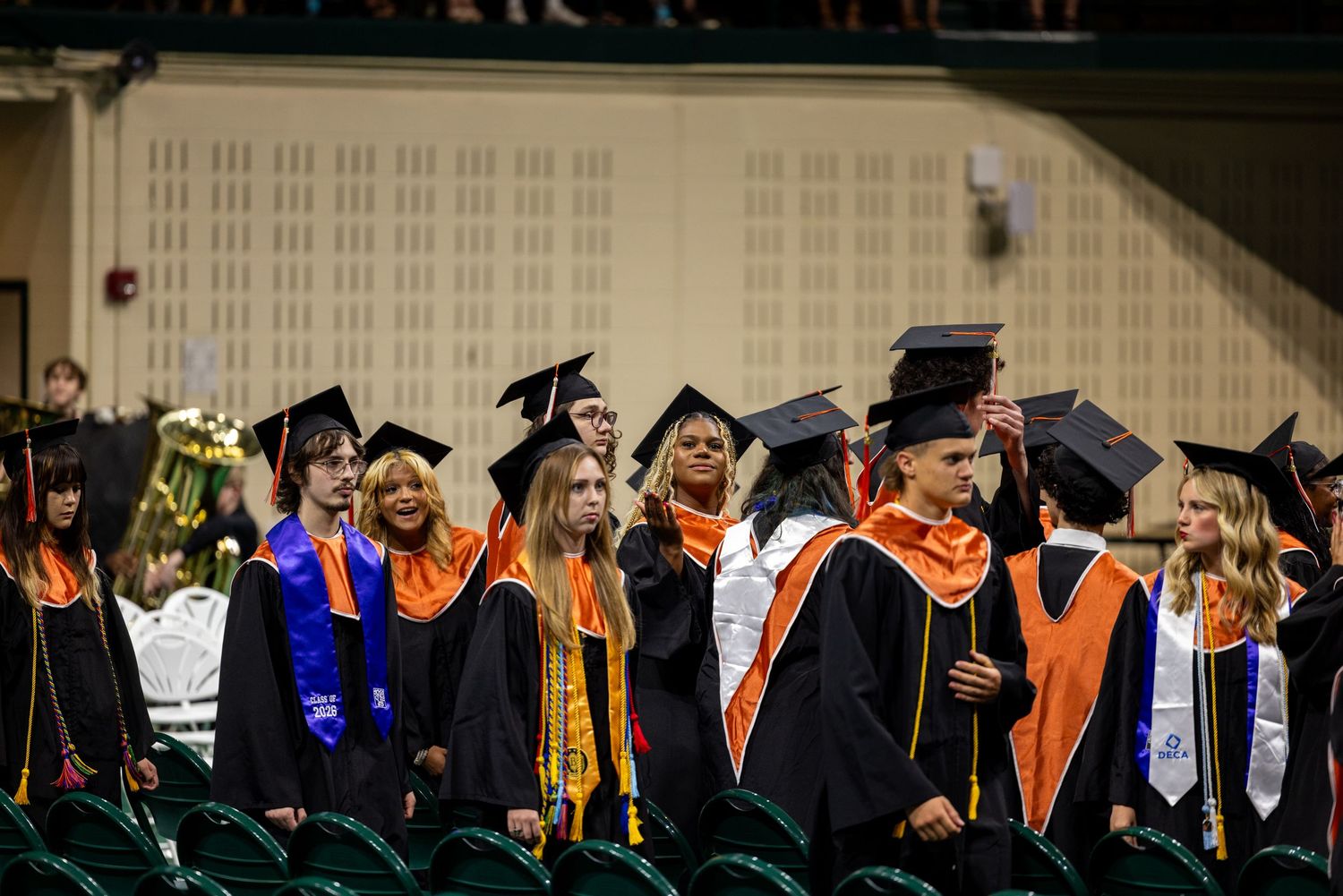 Rows of graduates in orange and black academic regalia stand at a college commencement ceremony.