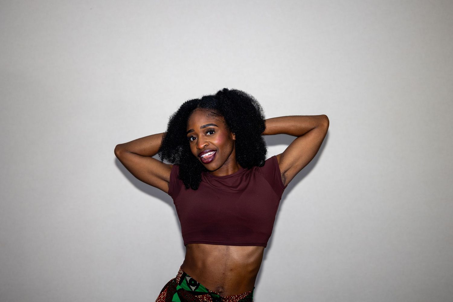 A fit Black woman with natural hair smiles in a burgundy crop top and colorful printed skirt.