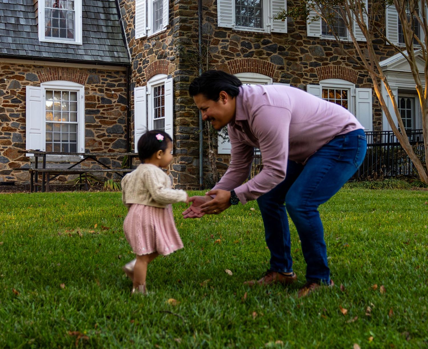 Adult bending down to hold hands with a toddler girl in pink dress on green lawn beside stone house.