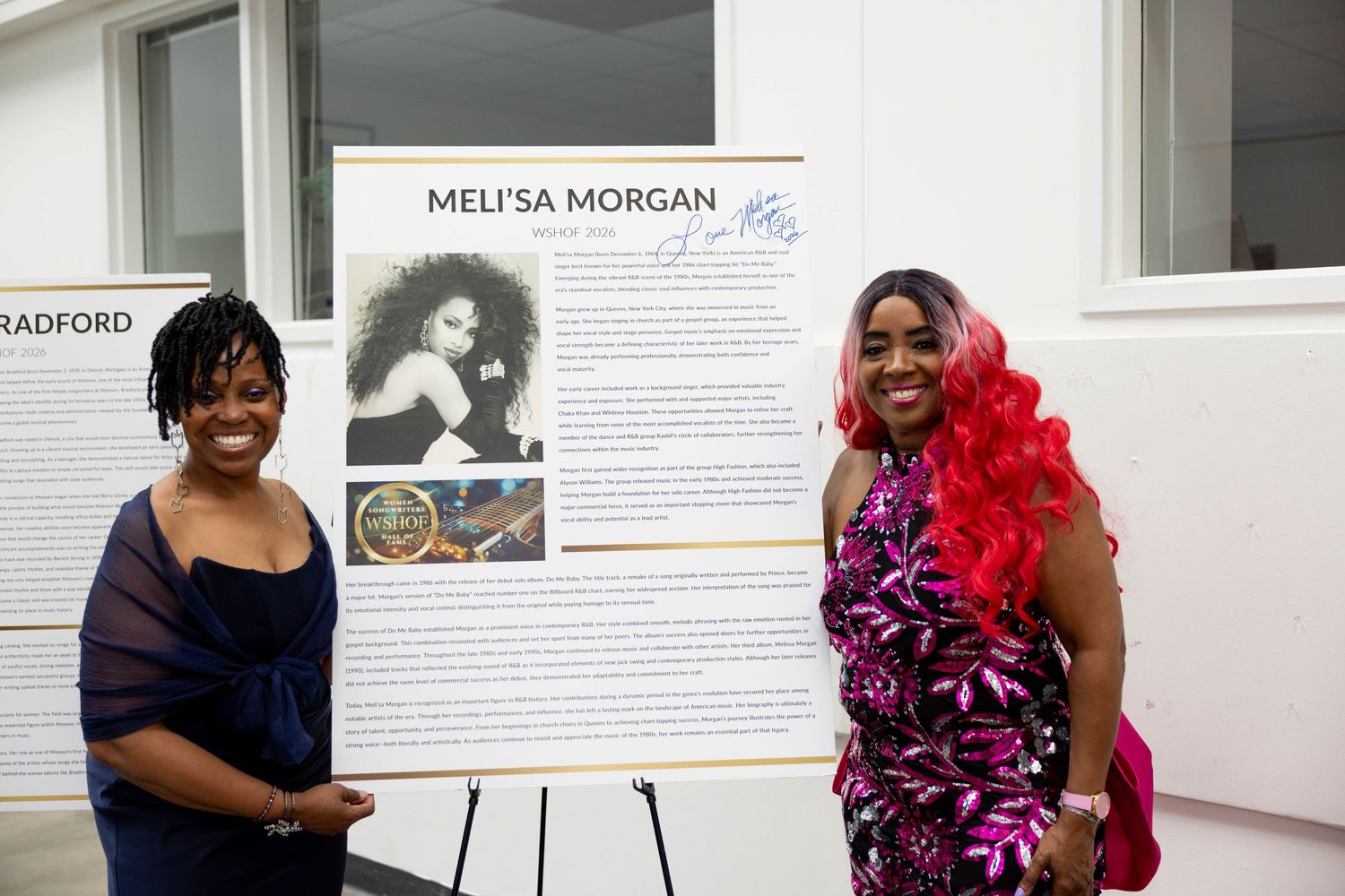 Two women posing beside a Meli'sa Morgan biography display board at an indoor event.