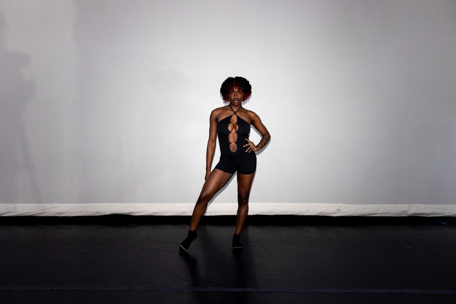 Woman in black cutout bodysuit strikes a confident pose against a light gray studio wall.